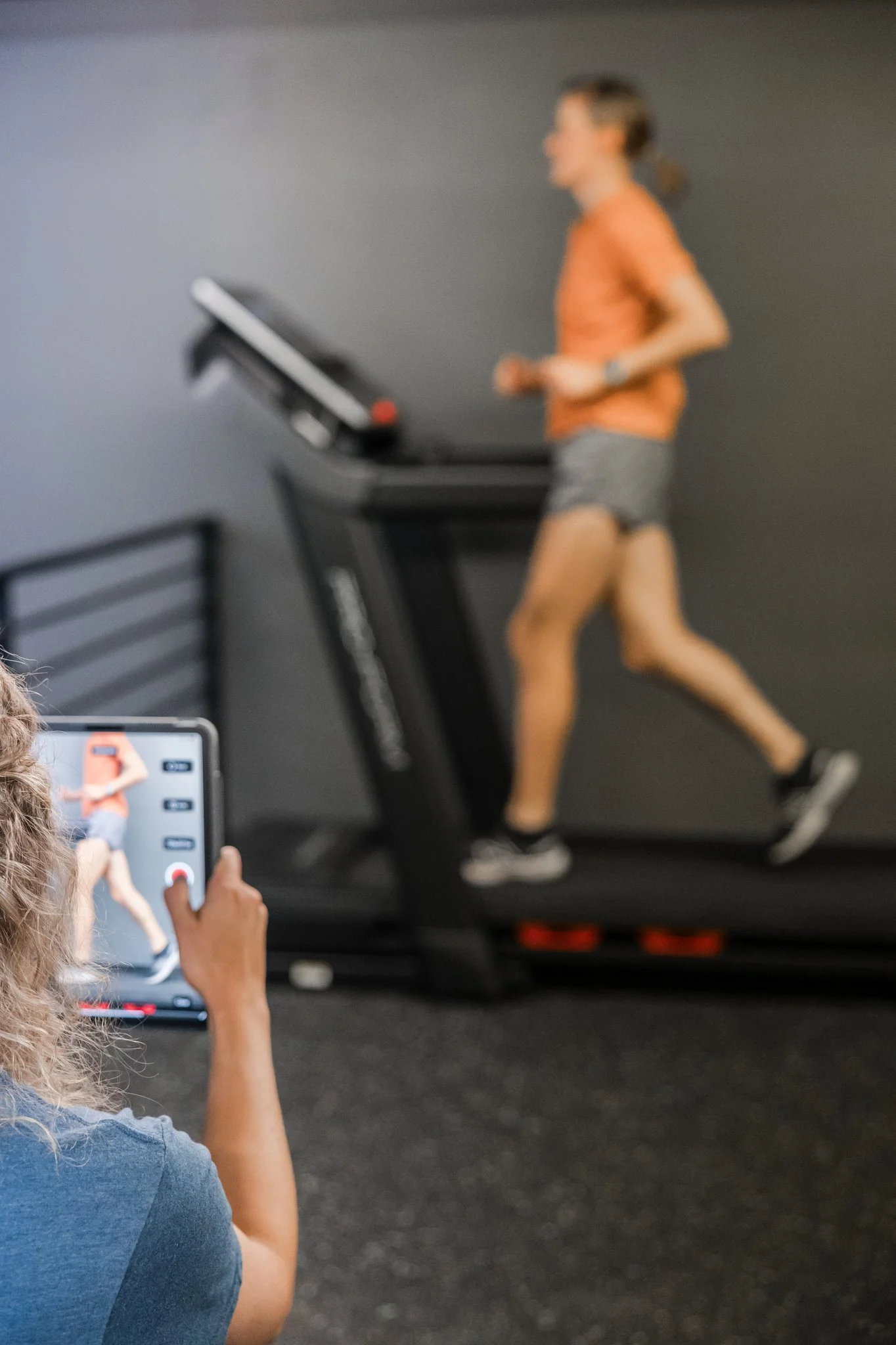 female patient in an orange shirt running on a treadmill while a physical therapist video tapes and analyzes gait
