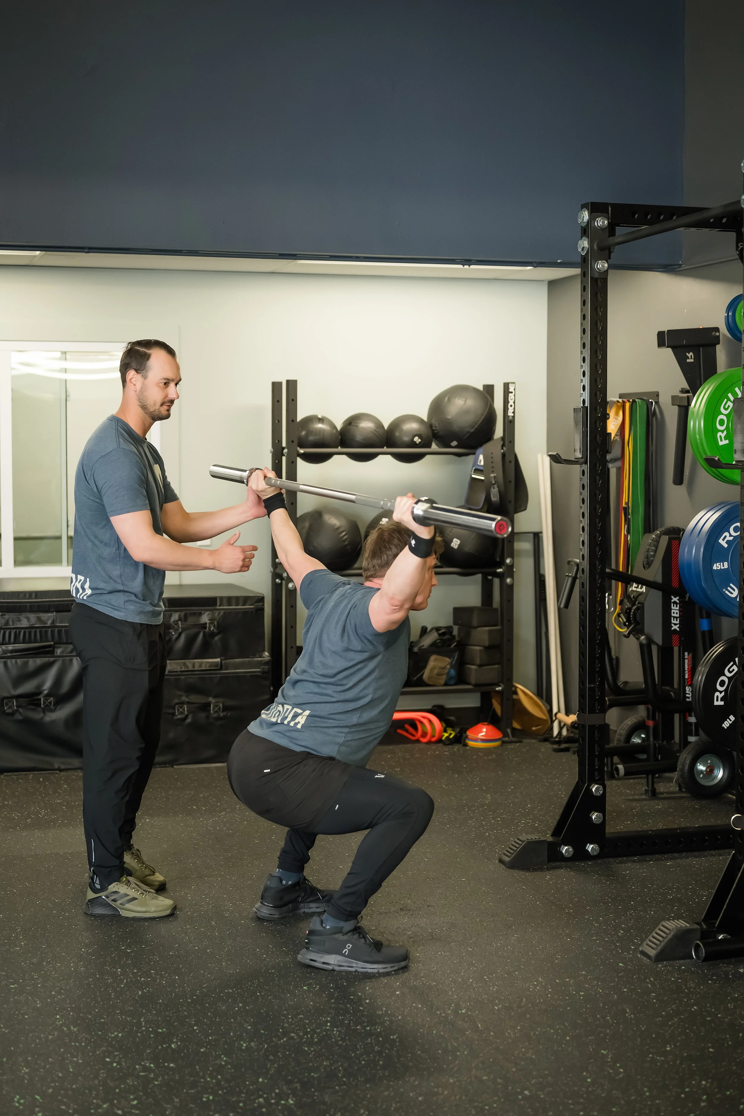 1 male helping another male with an overhead squat set up, both wearing blue shirts with medicine balls in the background