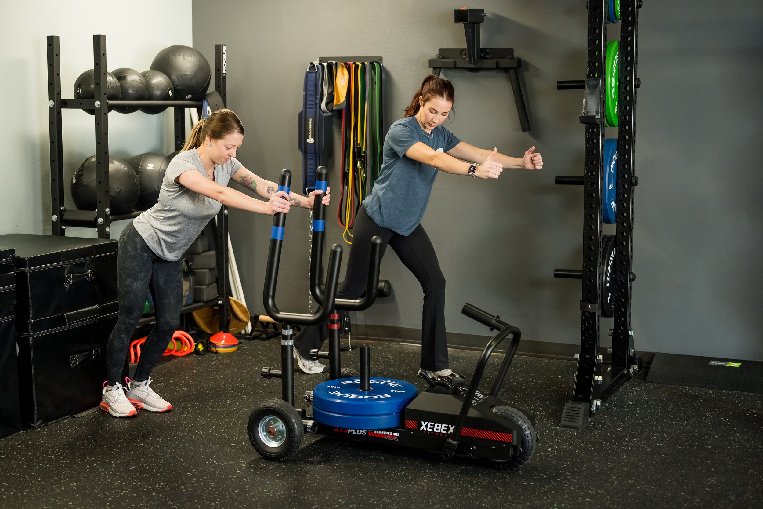 female in a grey shirt pushing a sled being assisted by a female therapist with bands and medicine balls in the background