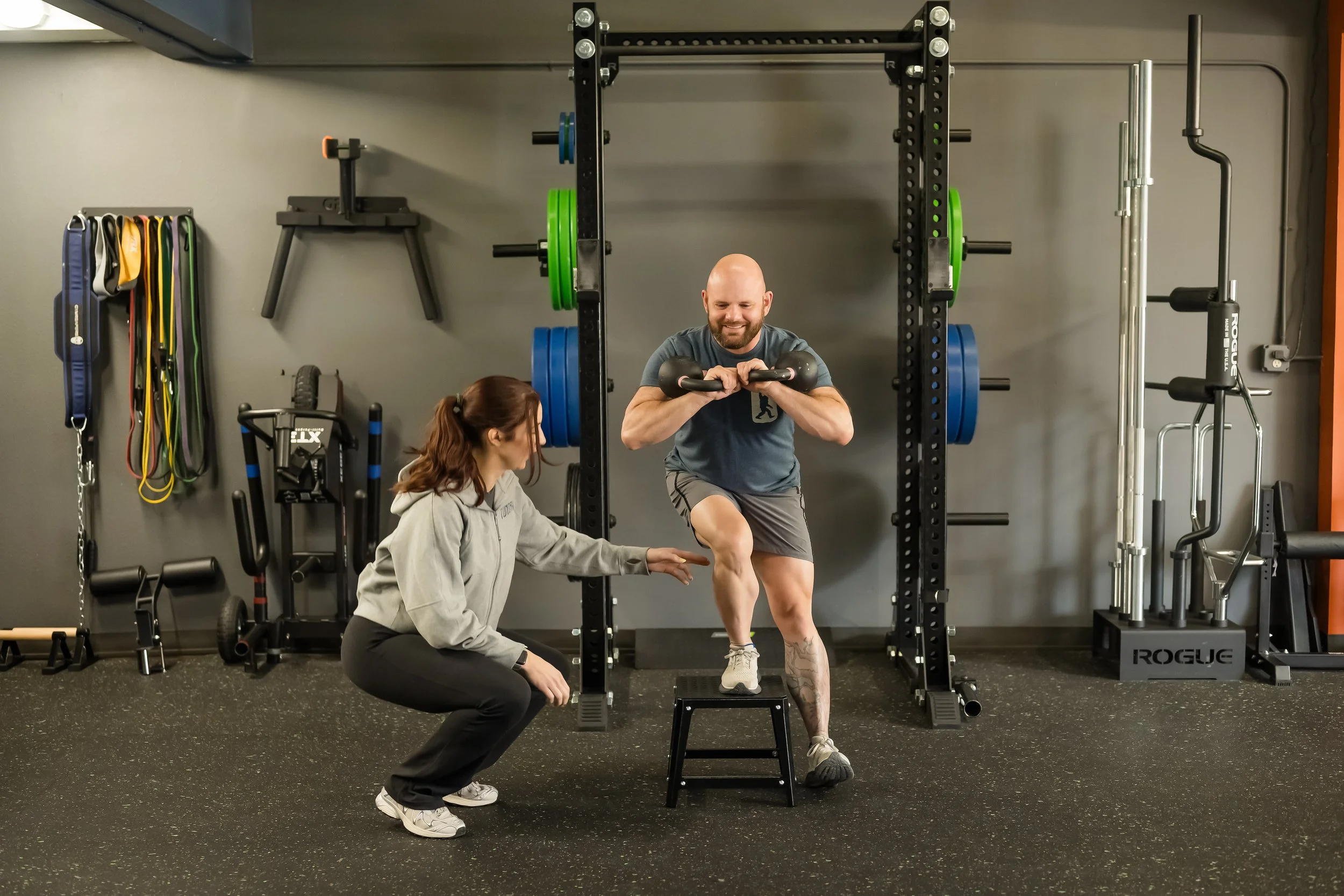 female in a grey sweater helping a male in a blue shirt completing a single leg squat with kettlebells with squat rack behind