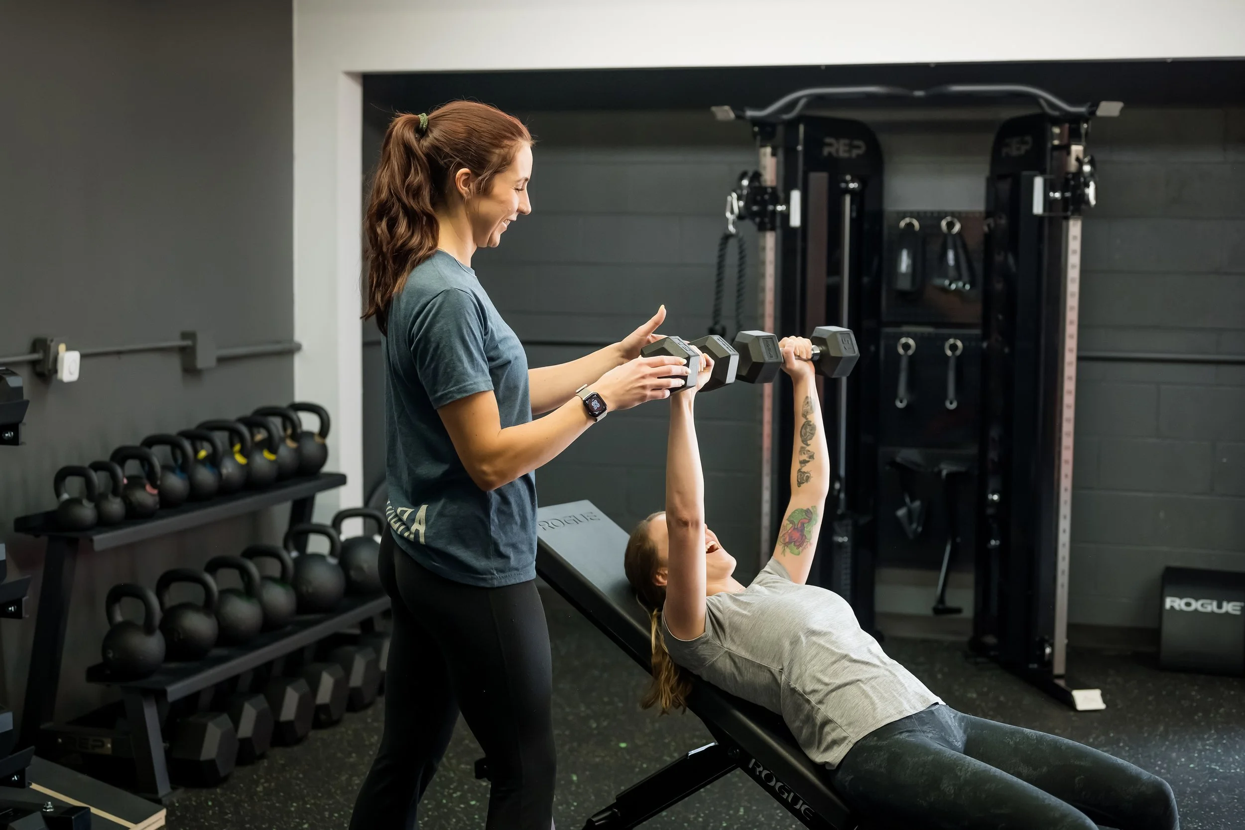 1 female in a blue shirt assisting a female in a grey shirt complete a dumbbell press in a gym with weights in the background