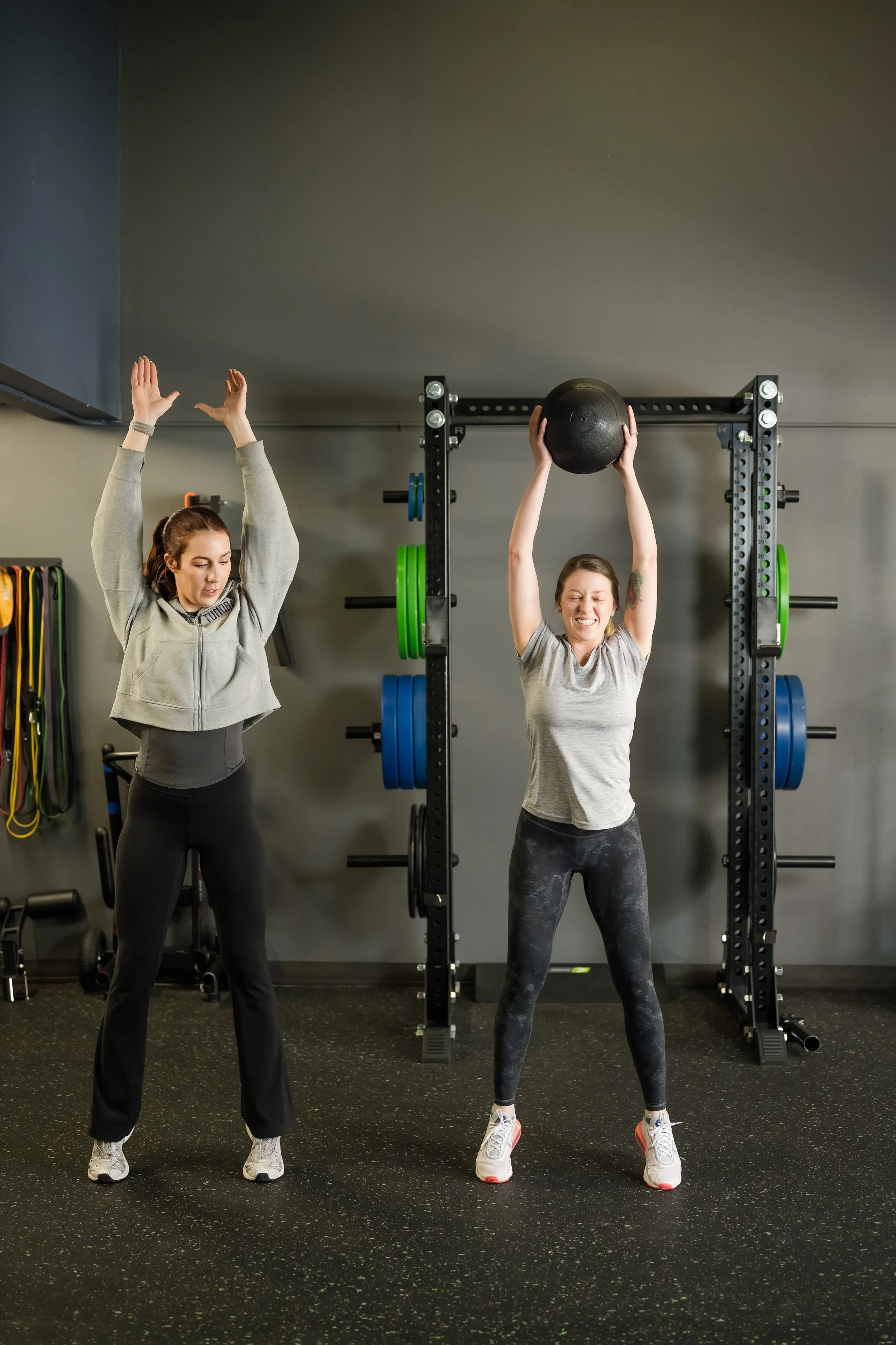 a woman in a grey sweater assisting a woman in a grey shirt with a medicine ball slam exercises