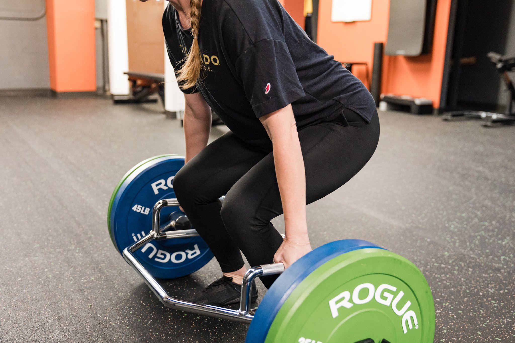 Person in black athletic clothing performing a deadlift with a barbell loaded with green and blue weight plates in a gym.