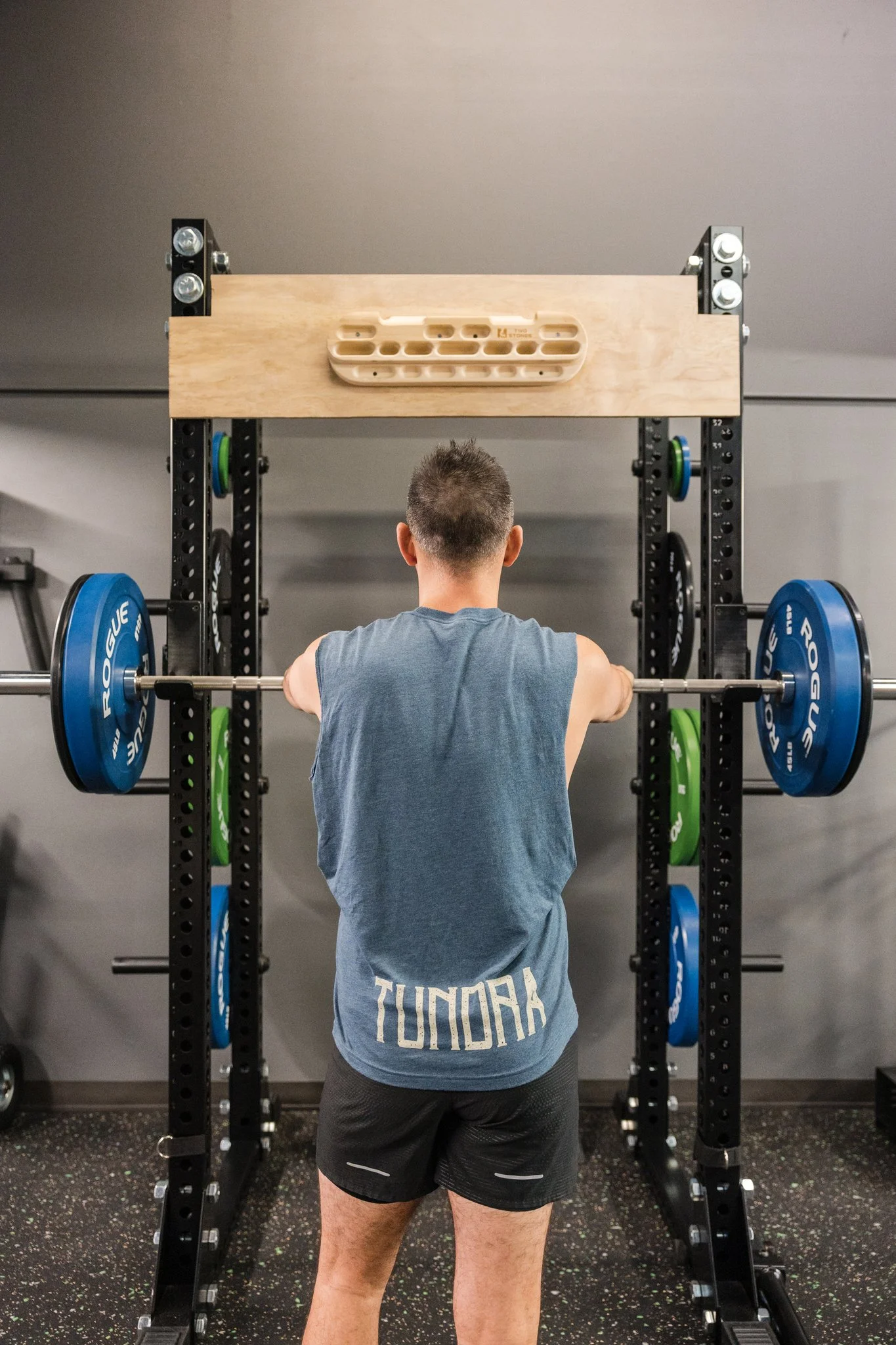 male patient wearing a blue tank top preparing to squat as part of his training program standing in front of a power rack with weight plates assembled attached