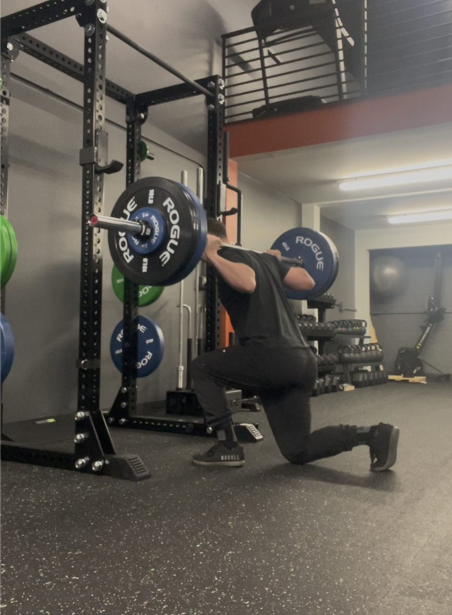 male weightlifter completing a barbell reverse lunge to full depth in a weight room in a power rack with weights stored on it
