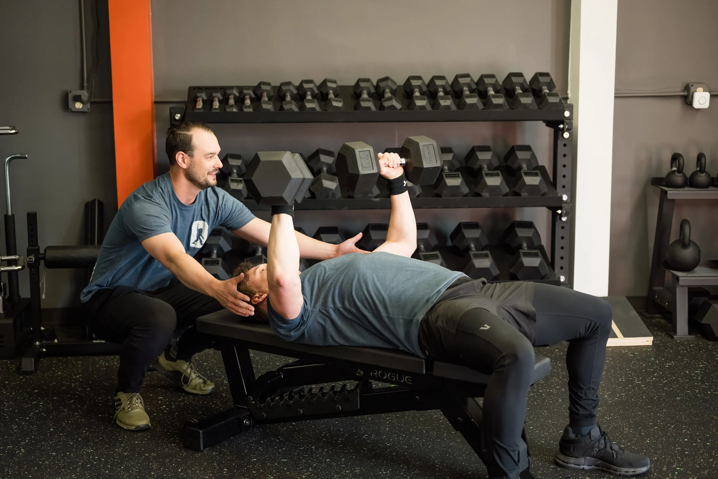 male patient in a blue tshirt being assisted by their provider in a blue tshirt lifting a dumbbell with a rack of dumbbells in the background