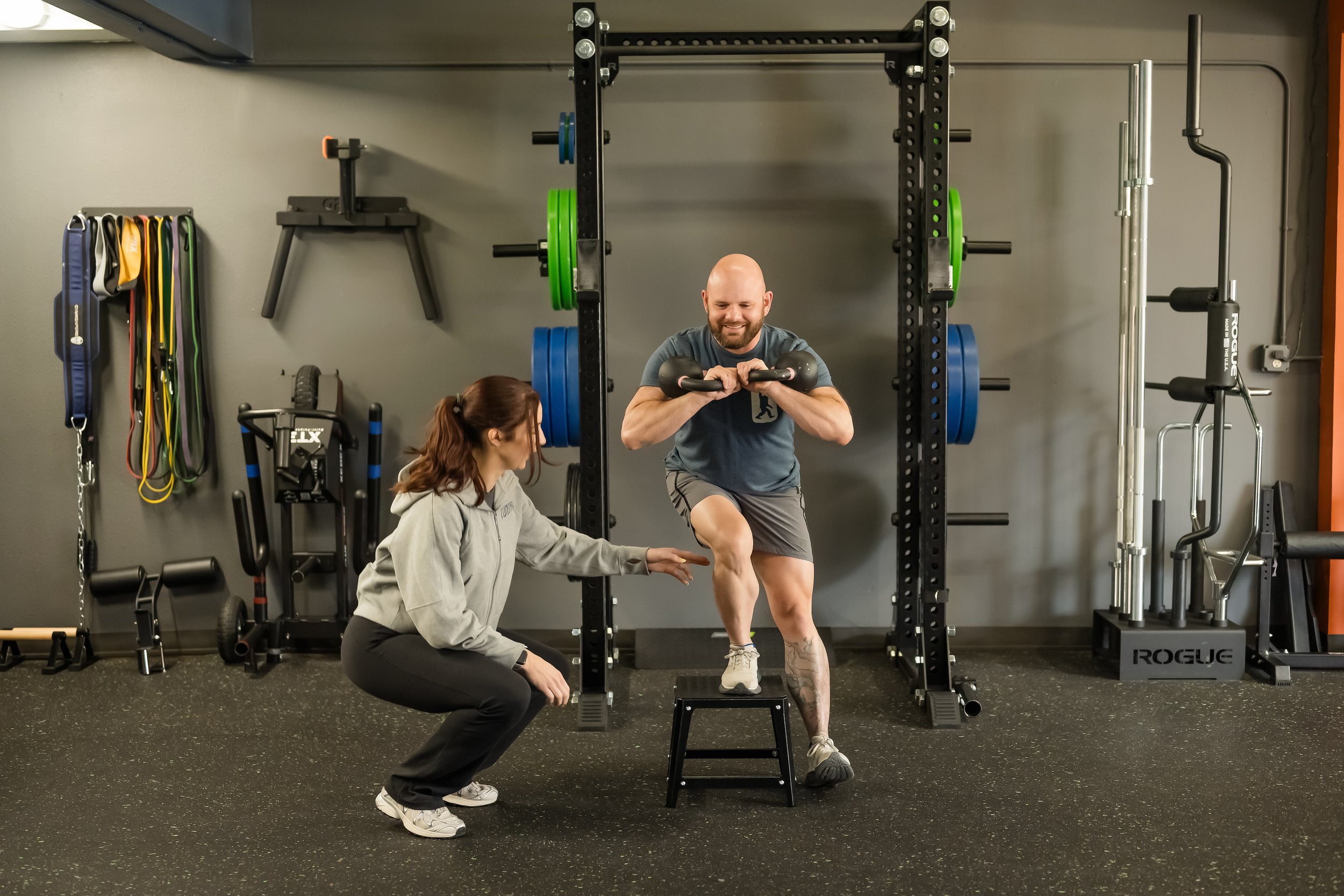 female in a grey sweater assisting a male in a blue shirt complete a single leg squat step down using kettlebells with a squat rack in the background
