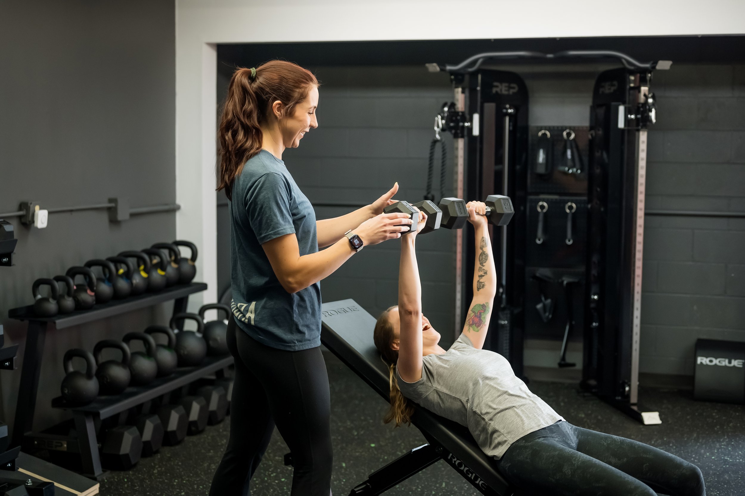 2 women lifting in the gym, one wearing a blue shirt, the other a grey shift lifting dumbbells