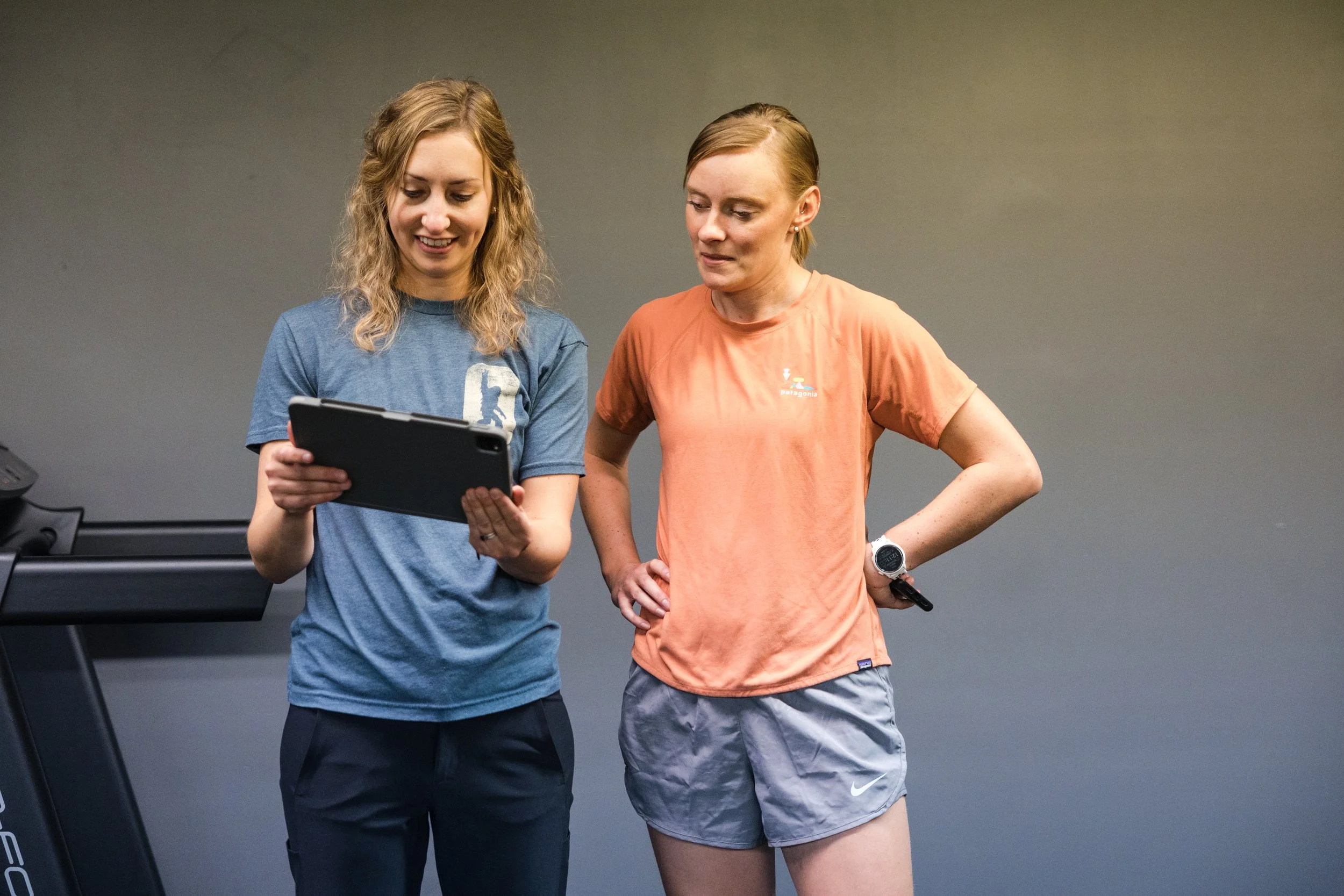 Two women in workout clothes looking at a tablet, in a fitness setting with a treadmill.