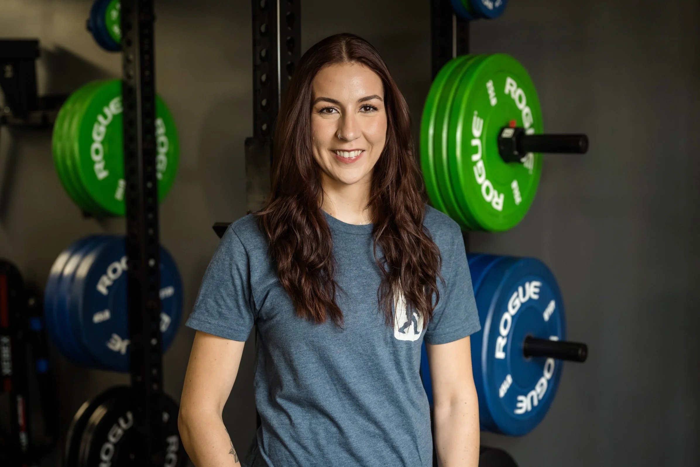 A smiling woman with brown hair, wearing a blue t-shirt with a gym logo, is in a gym with weightlifting equipment in the background.