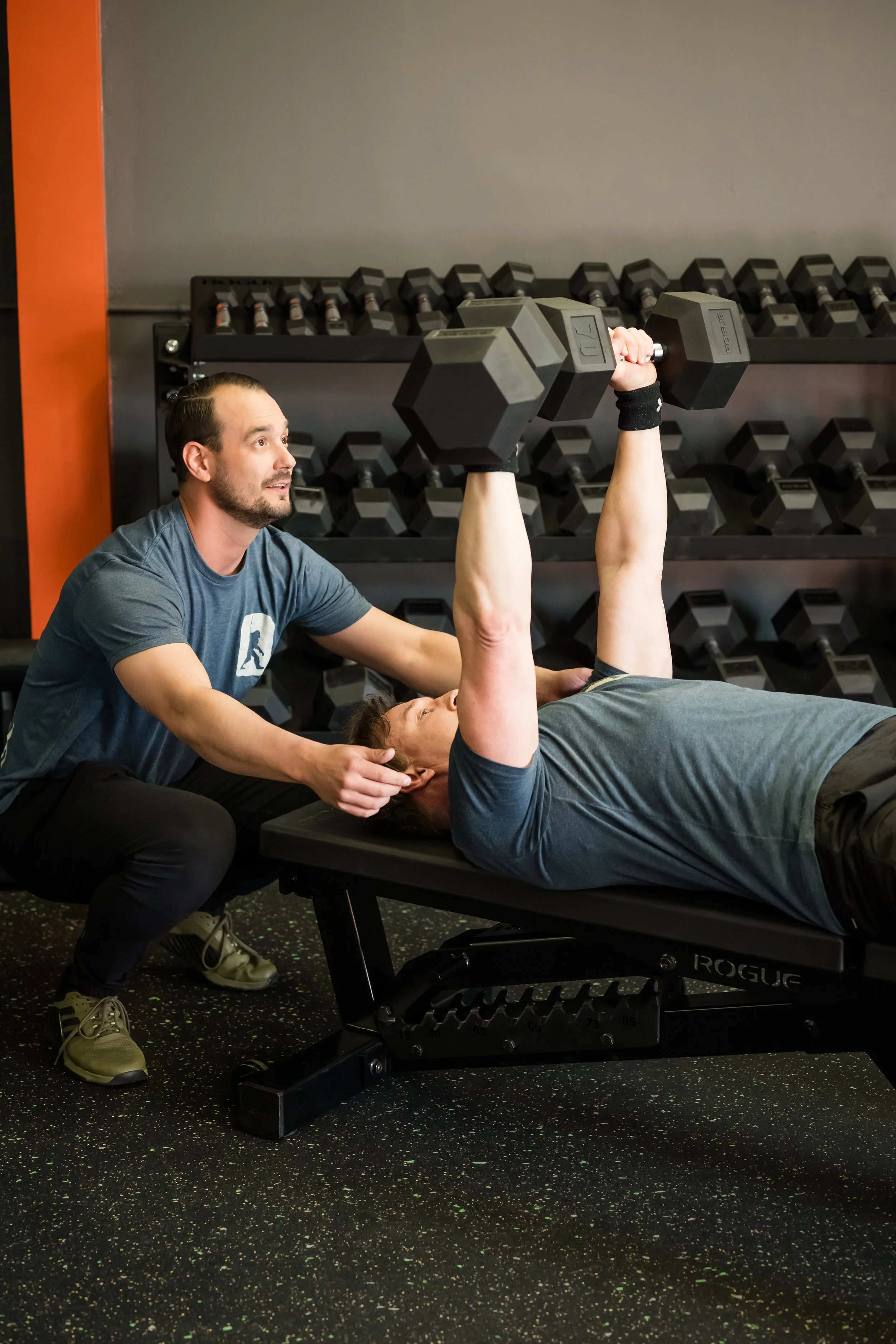 two males wearing blue tshirts bench pressing with dumbbells
