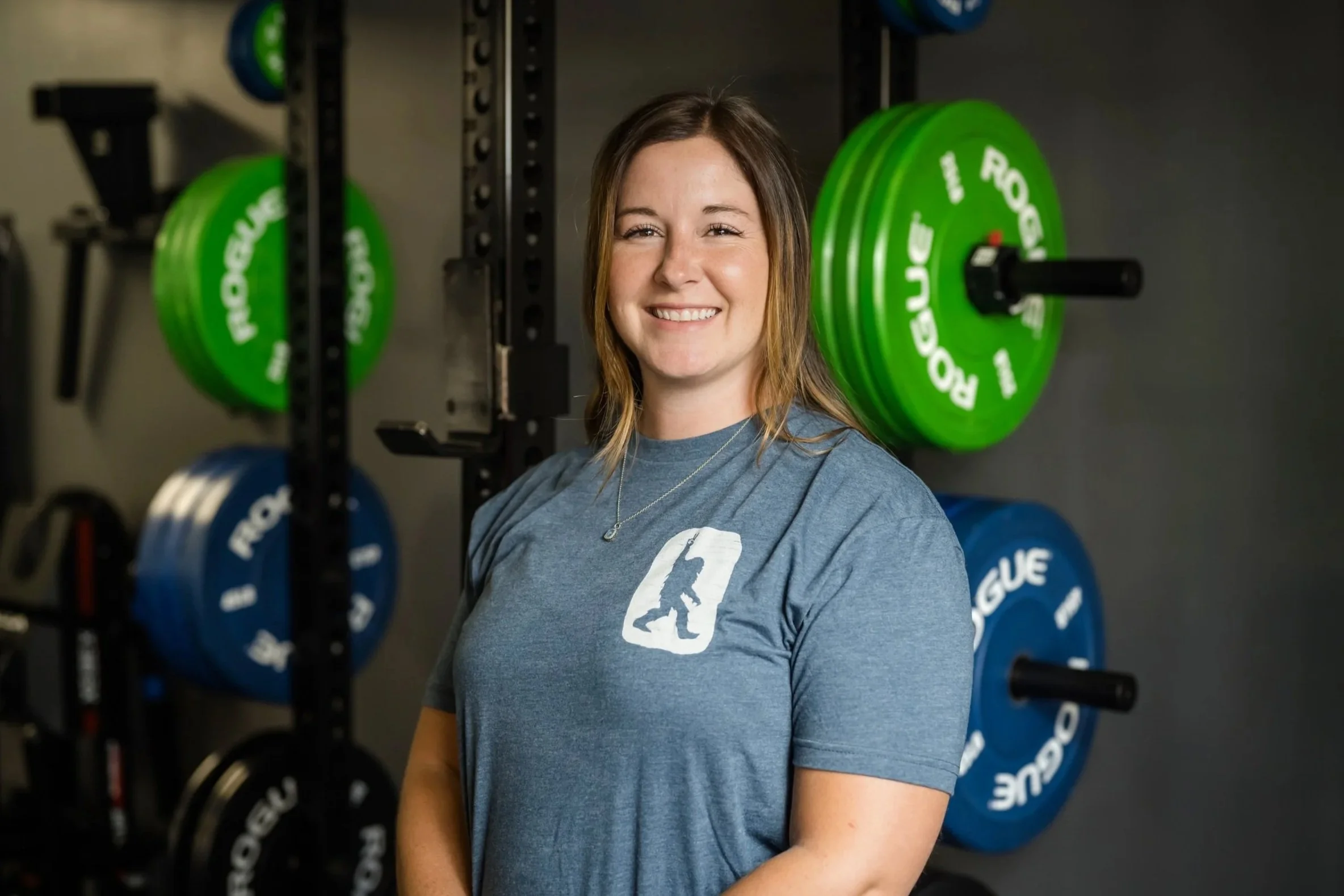 A smiling woman with brown hair, wearing a blue t-shirt with a gym logo, is in a gym with weightlifting equipment in the background.