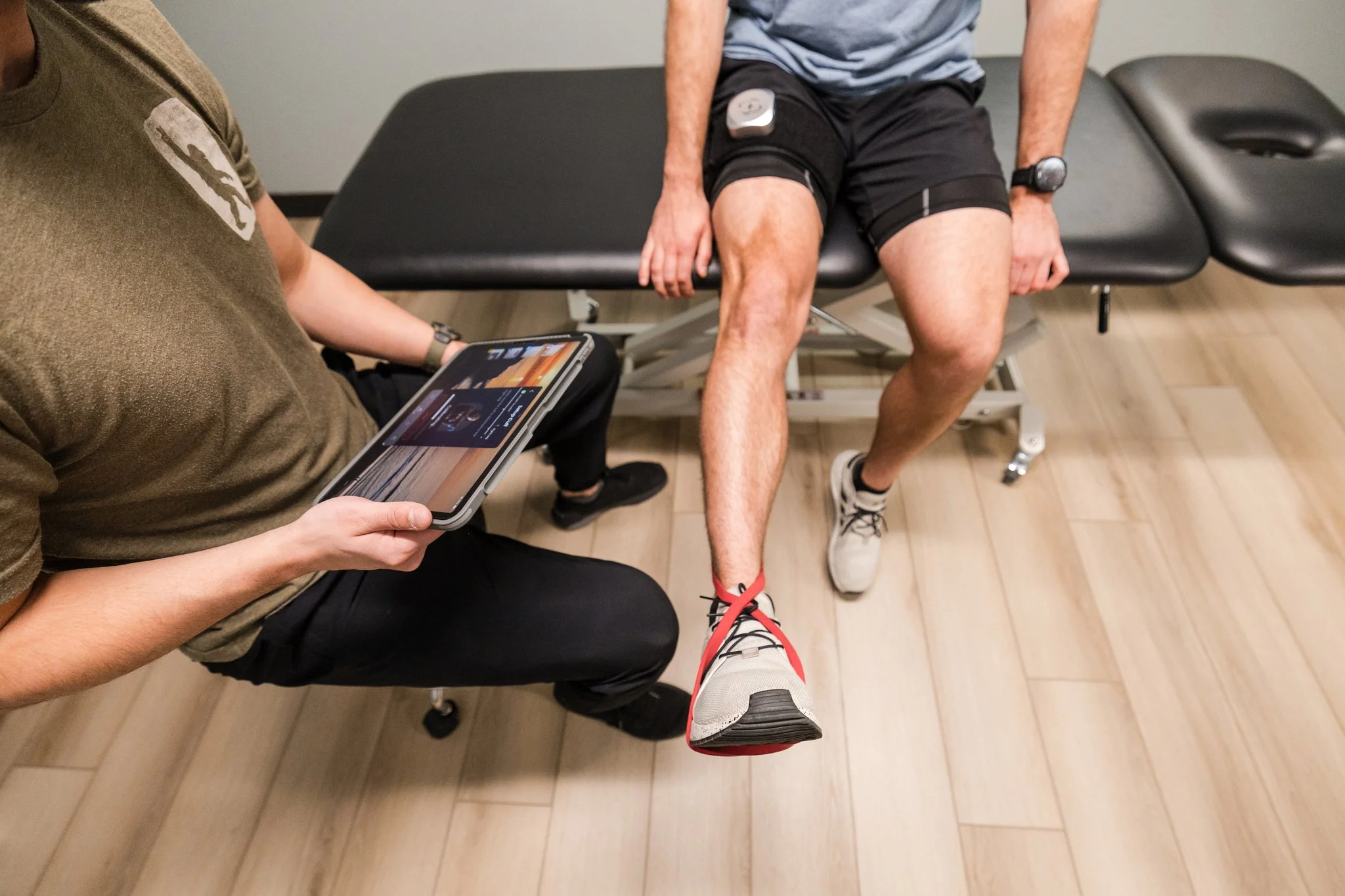 male athlete in a blue shirt using blood flow restriction therapy along with another male therapist in a green shirt holding an ipad to provide visual feedback