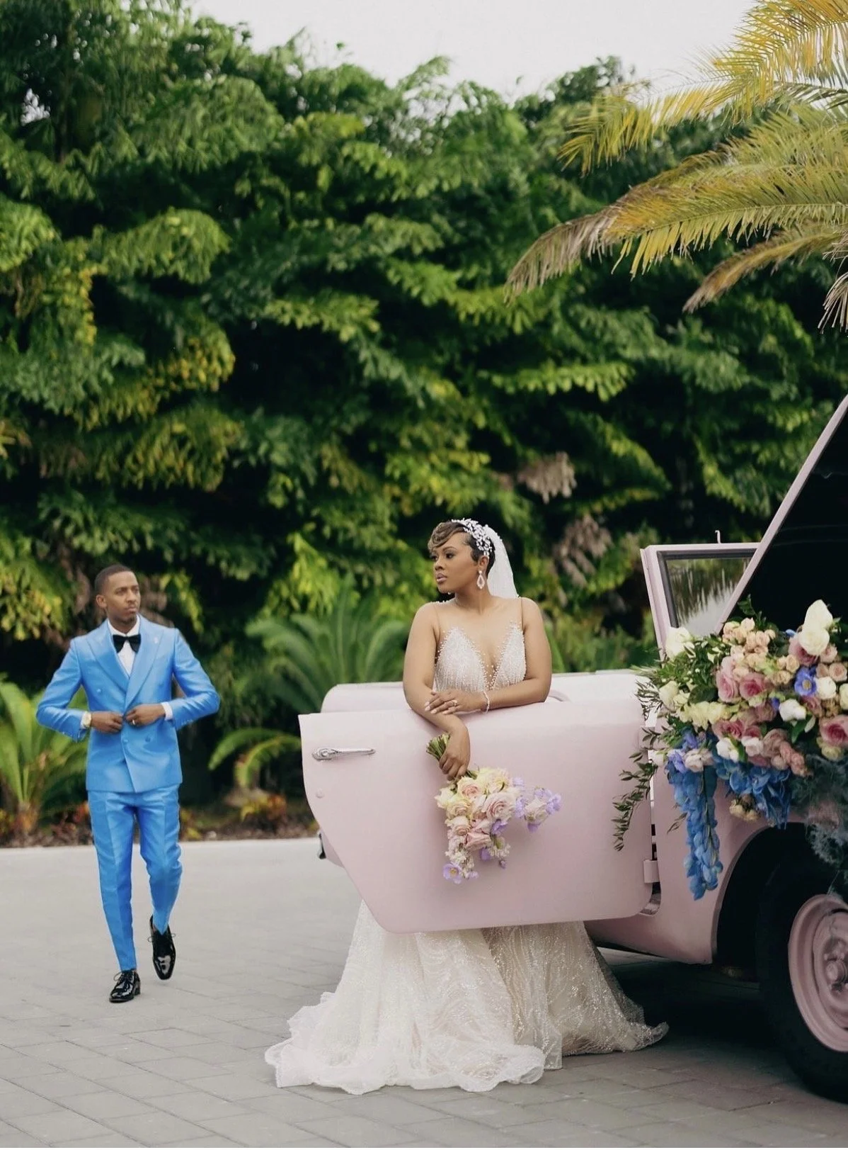 A couple on their wedding day in front of greenery. The bride is standing and leaning over an opened pink door of a car and the groom is walking up behind her.