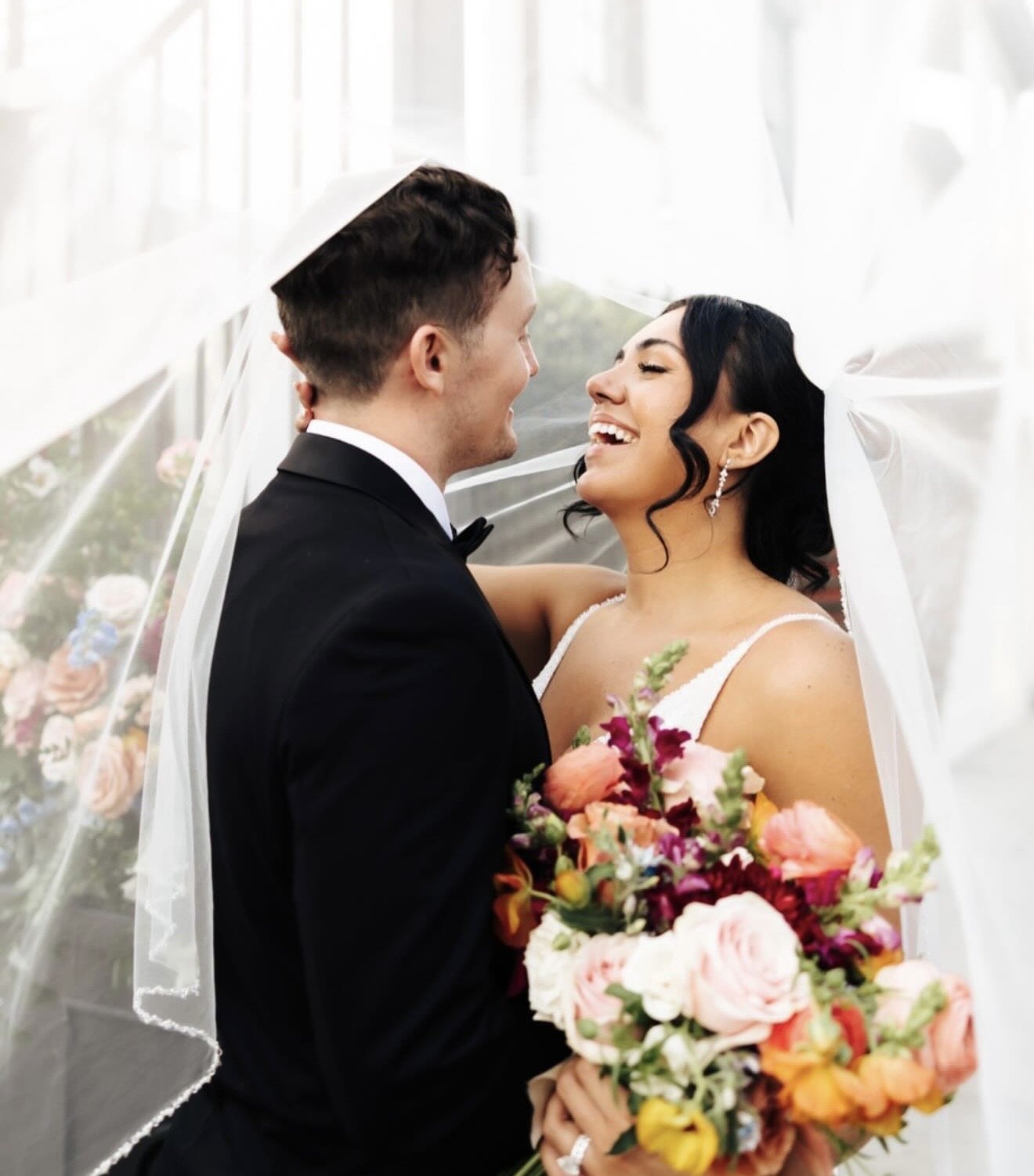 A bride and groom smiling at each other under the bride's veil as she holds a color bouquet.