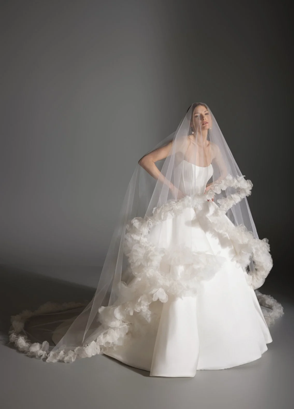 A woman in a wedding dress with a veil standing against a plain gray background.