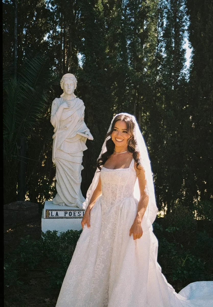 A bride outside standing in front of a statue and greenery smiling.