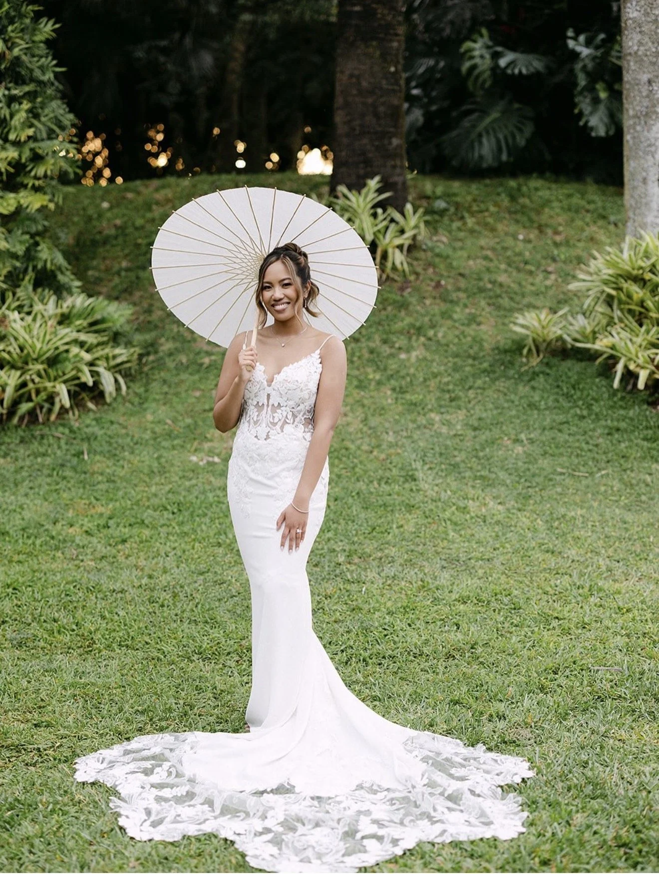 A bride holding a white umbrella in front of greenery.