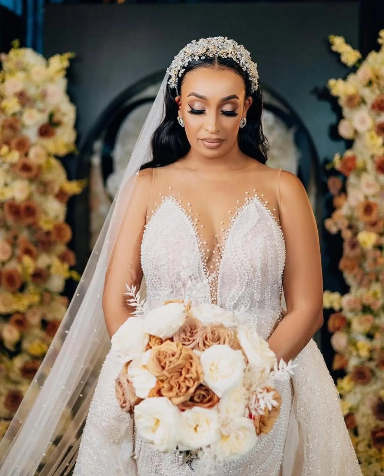 A bride standing in front of an arch of flowers holding a bouquet and looking down.