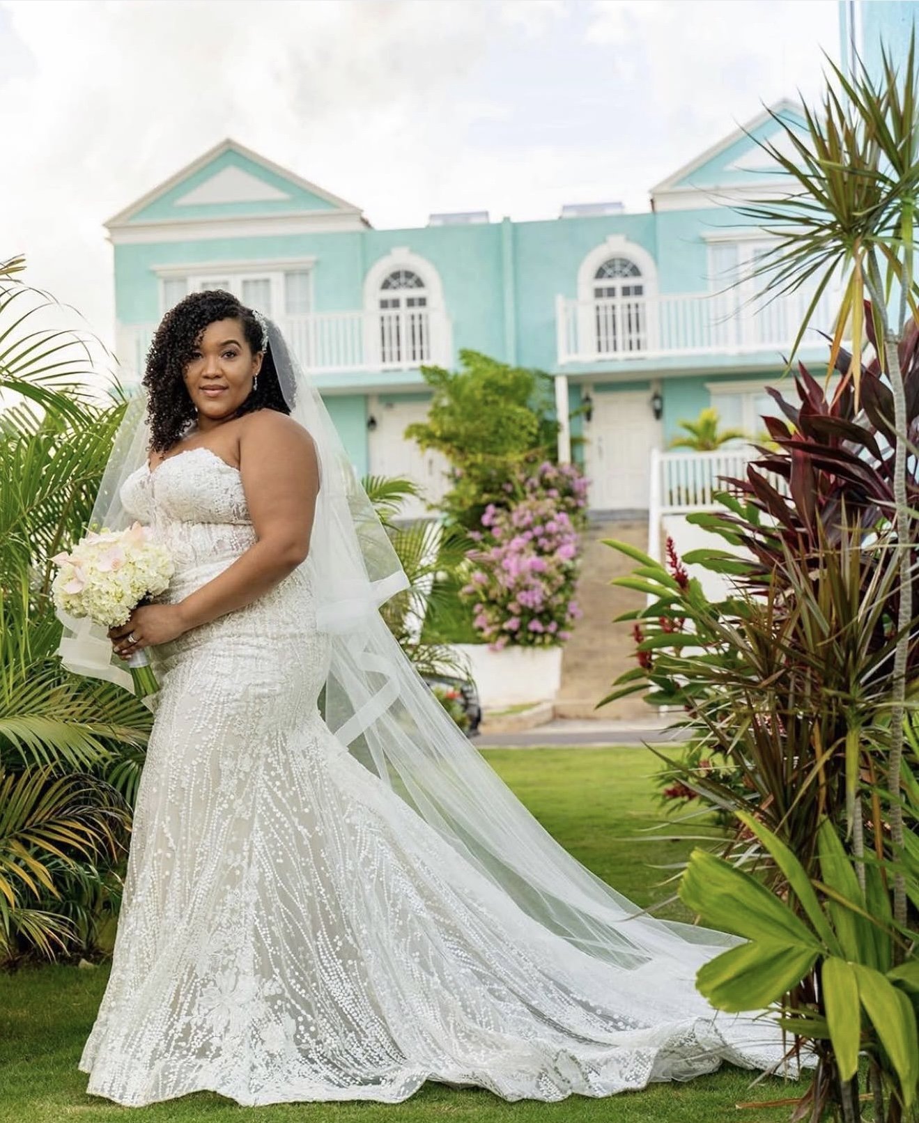 A bride in a white wedding dress holding a bouquet, standing outdoors in front of a pastel blue house with lush greenery and pink flowers.