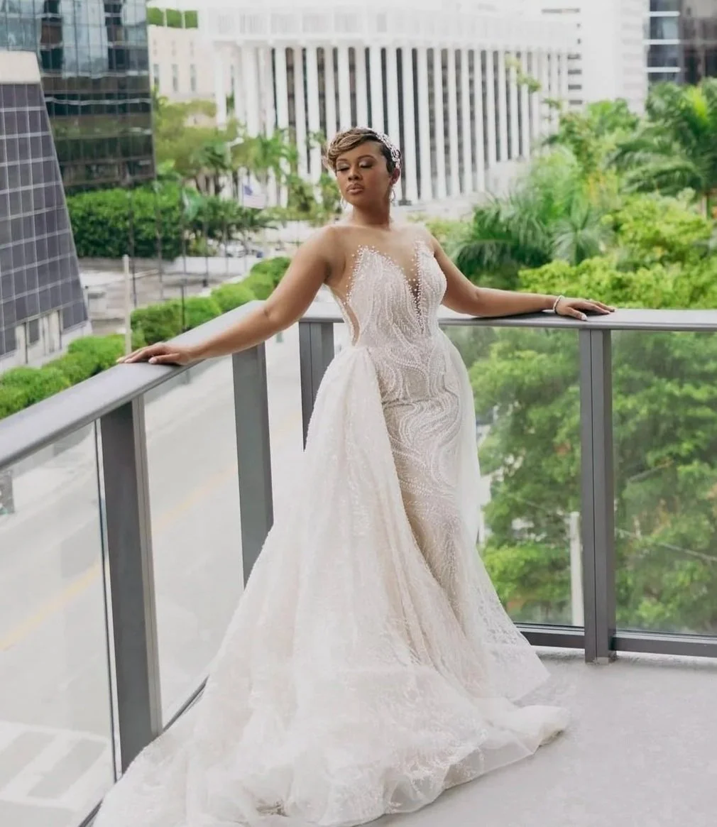 A woman in a wedding dress standing on a balcony with city buildings and trees in the background.