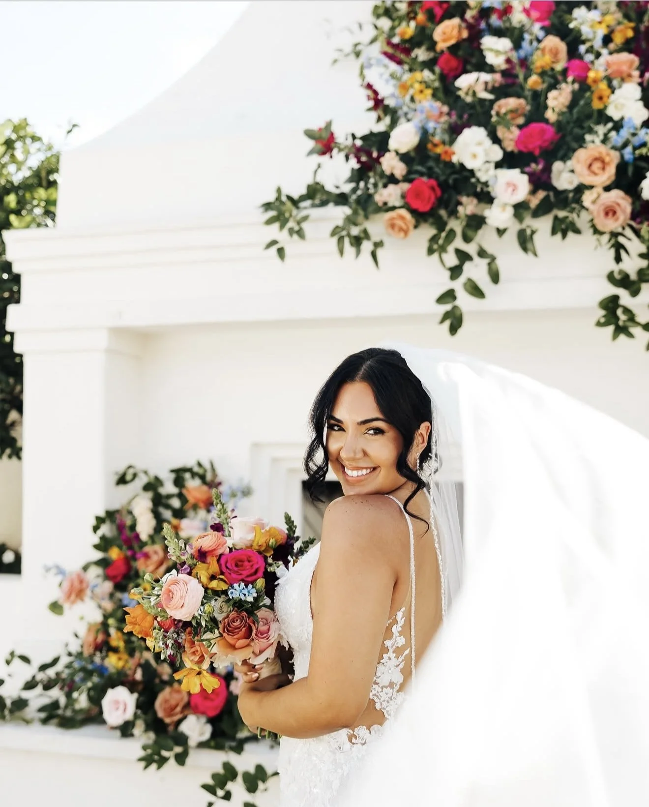 A smiling bride in a white wedding dress holding a colorful bouquet of flowers surrounded by more flowers in the background.