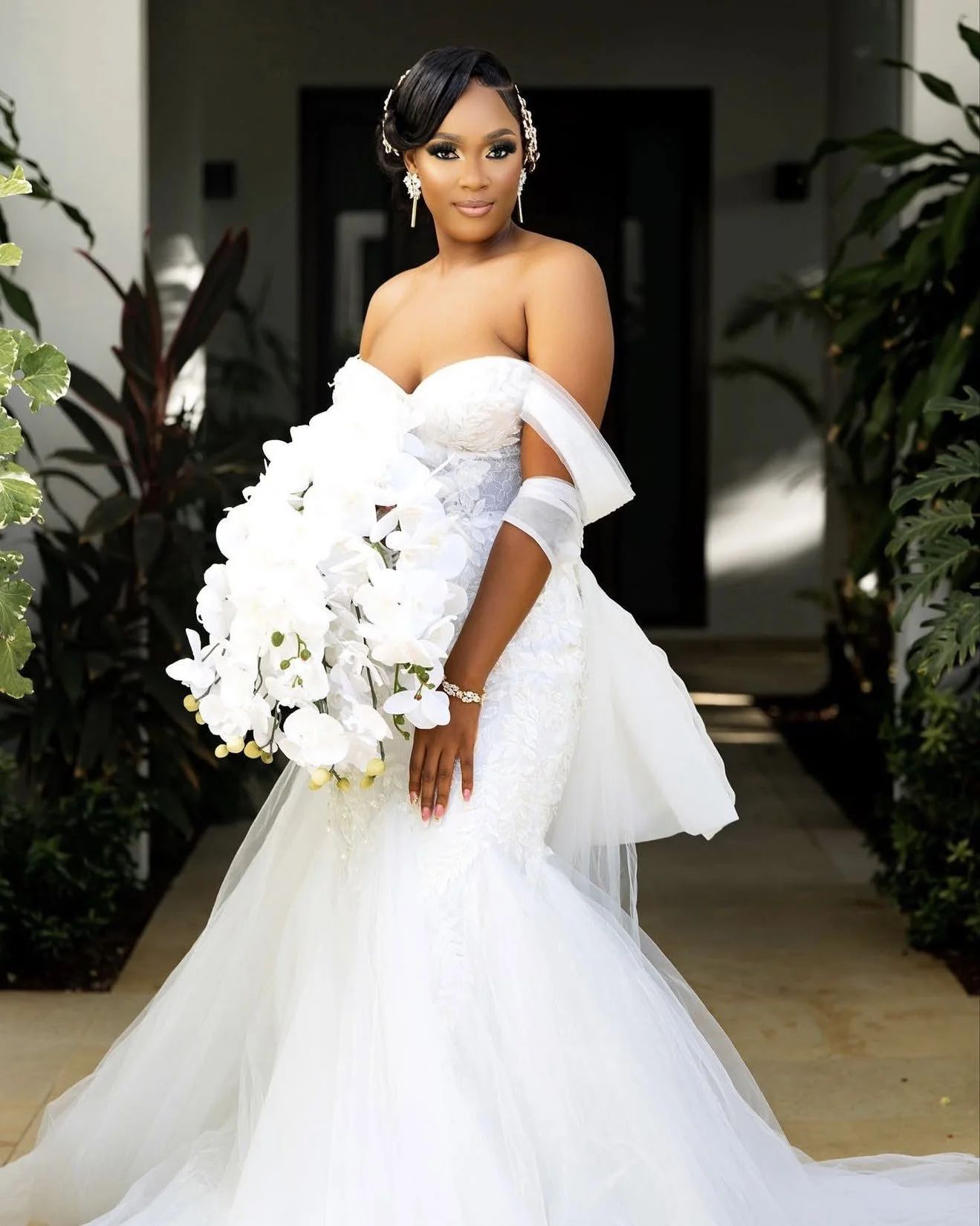 A bride in a white wedding gown holding a bouquet of white orchids, standing indoors surrounded by green plants.