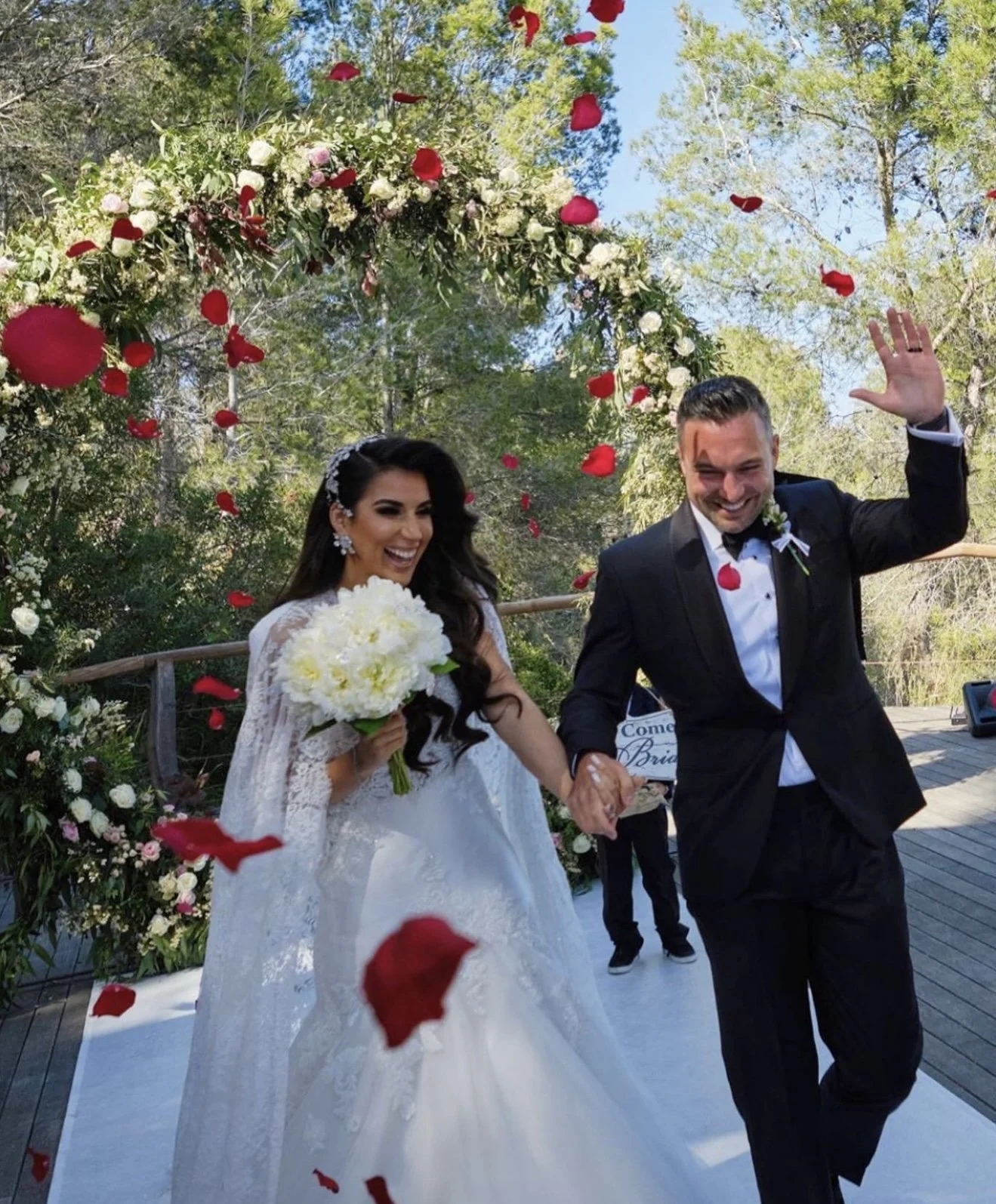 A newlywed couple is walking under a floral archway outdoors, holding hands, smiling, as red and white rose petals fall around them during their wedding celebration.