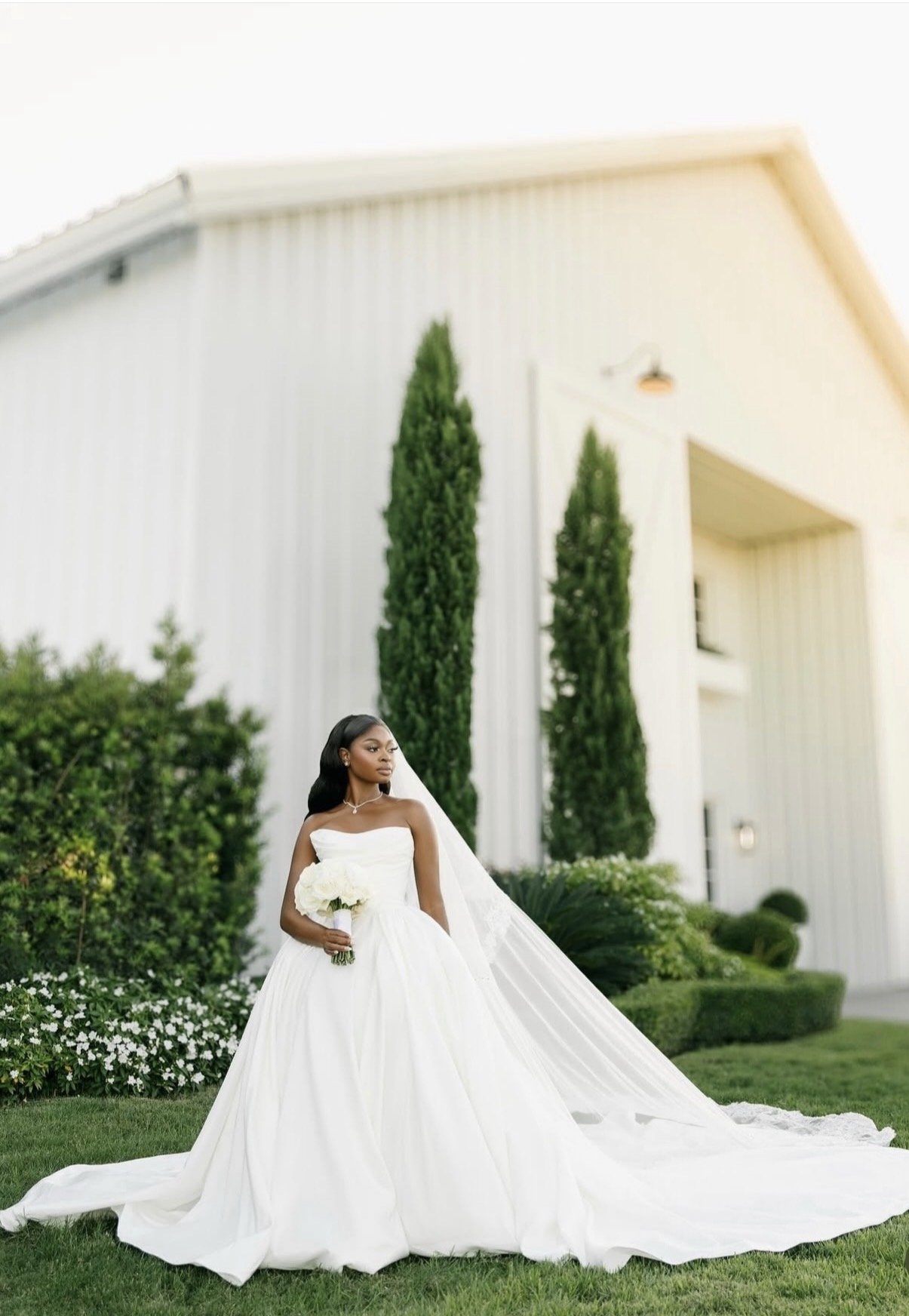 A bride outdoors in front of green bushes and a white building.