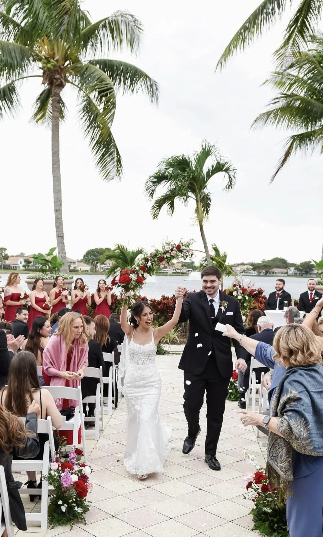 A happy couple walking down the aisle after getting married with their wedding party and guests looking on. They are outside with palm trees and water in the background.