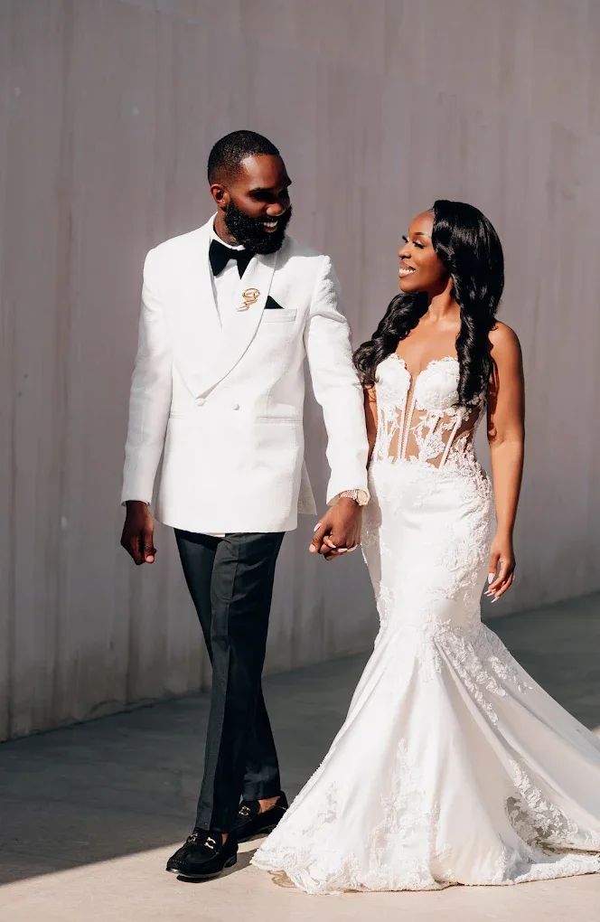A bride and groom walking hand in hand indoors, with the bride wearing a white lace wedding dress and the groom in a white tuxedo jacket with black pants.