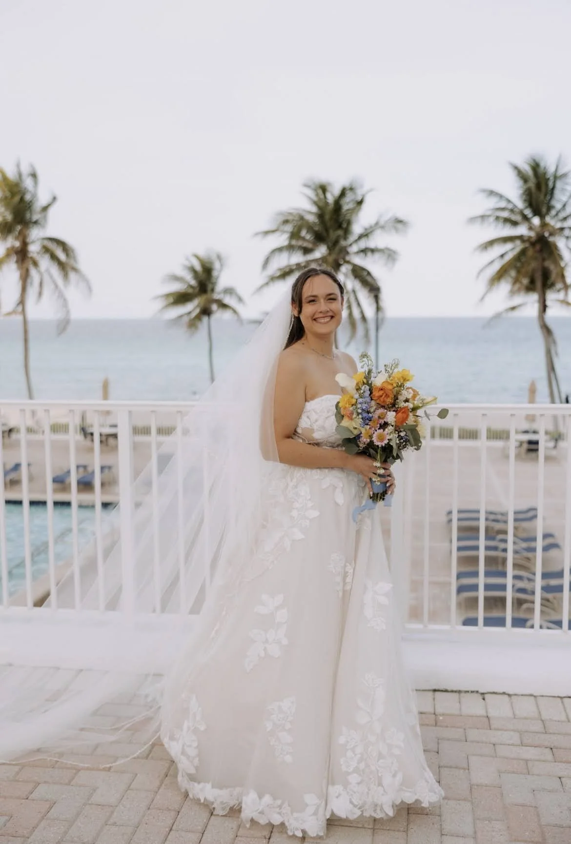 A bride outside standing on a balcony holding a bouquet with palm trees and water in the background.