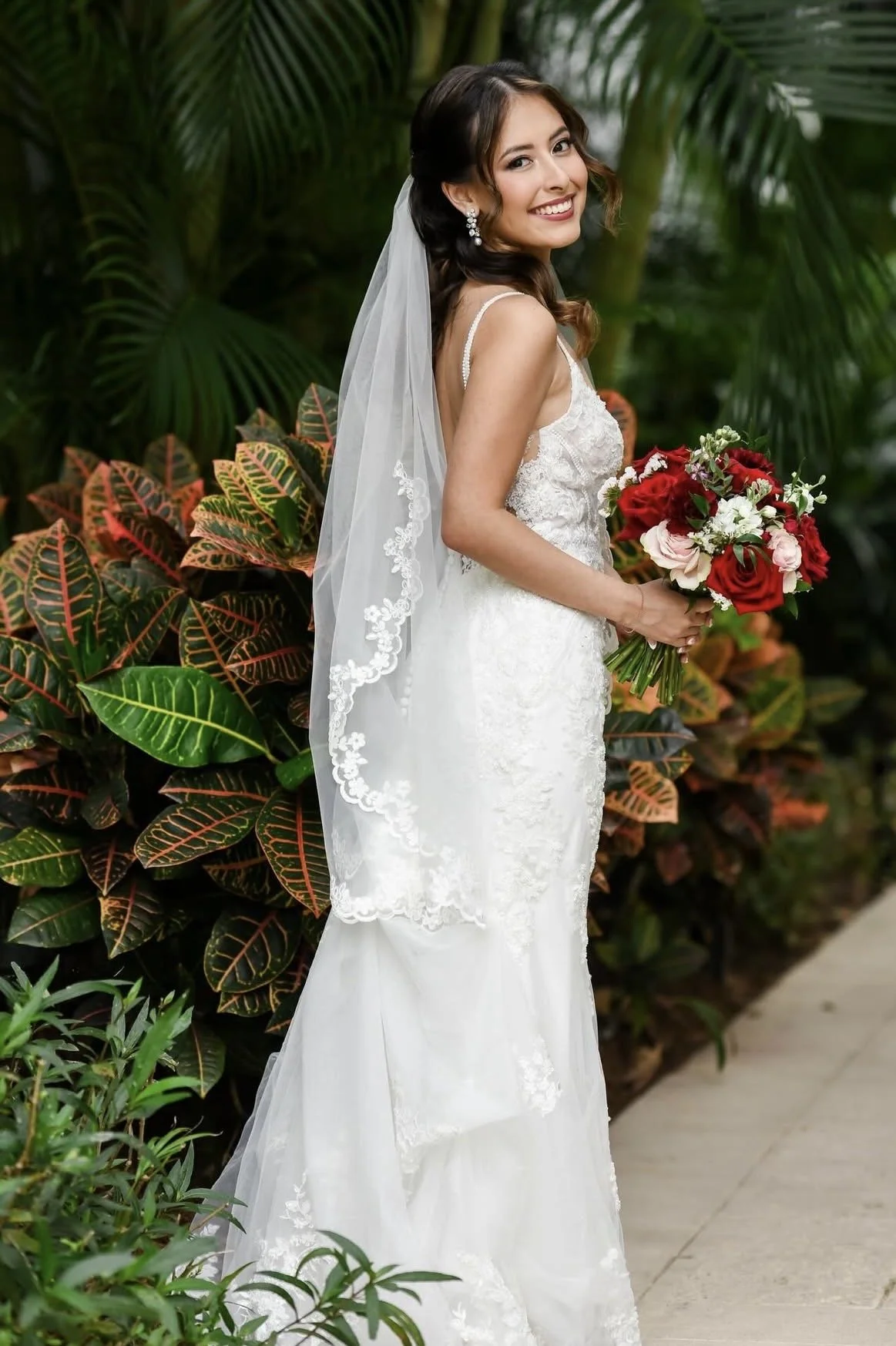 A bride smiling outside looking over her shoulder holding a bouquet in front of greenery.