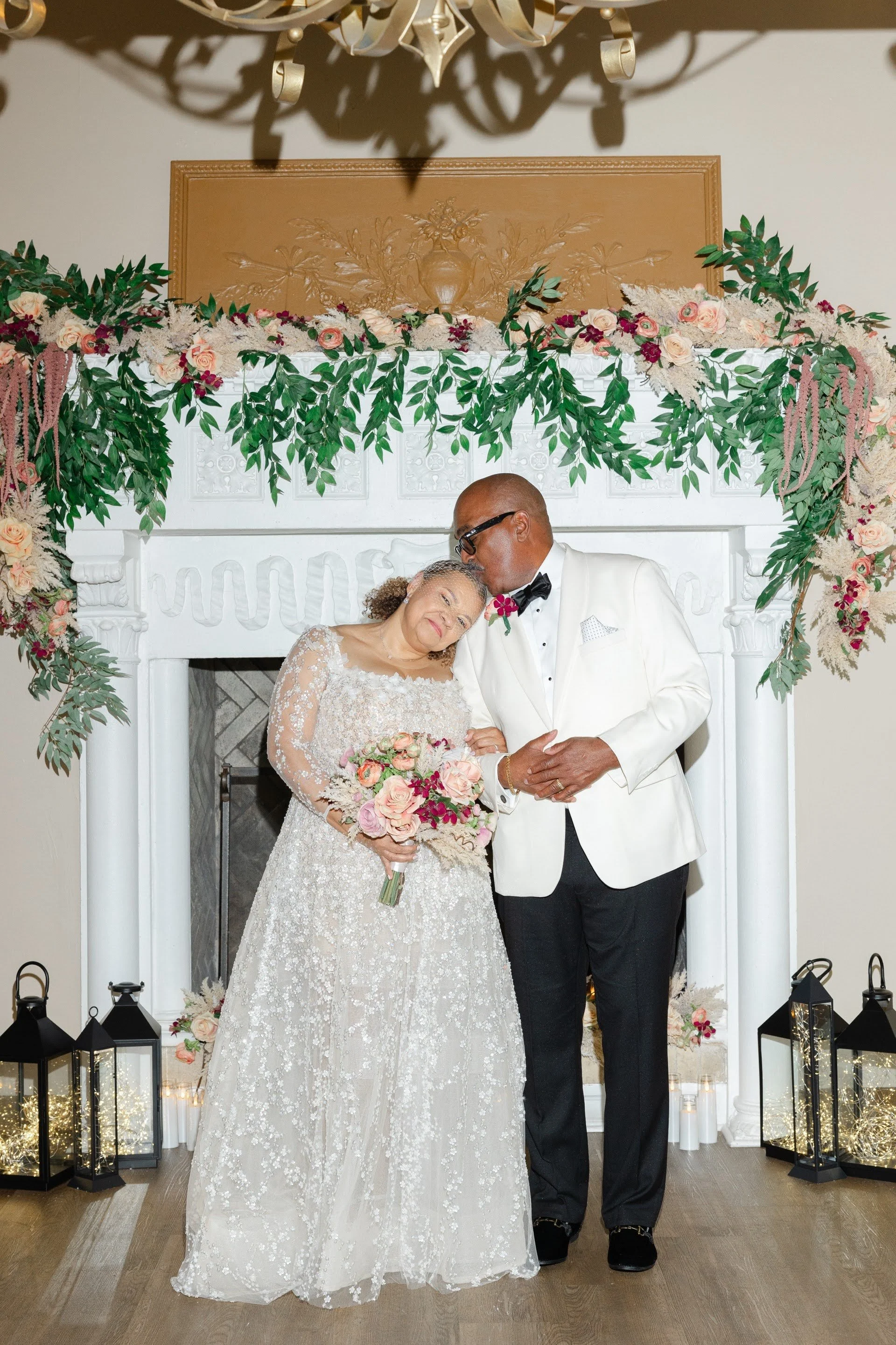 A bride and groom standing in front of a fireplace with flowers above it. The groom is kissing the bride on her head.