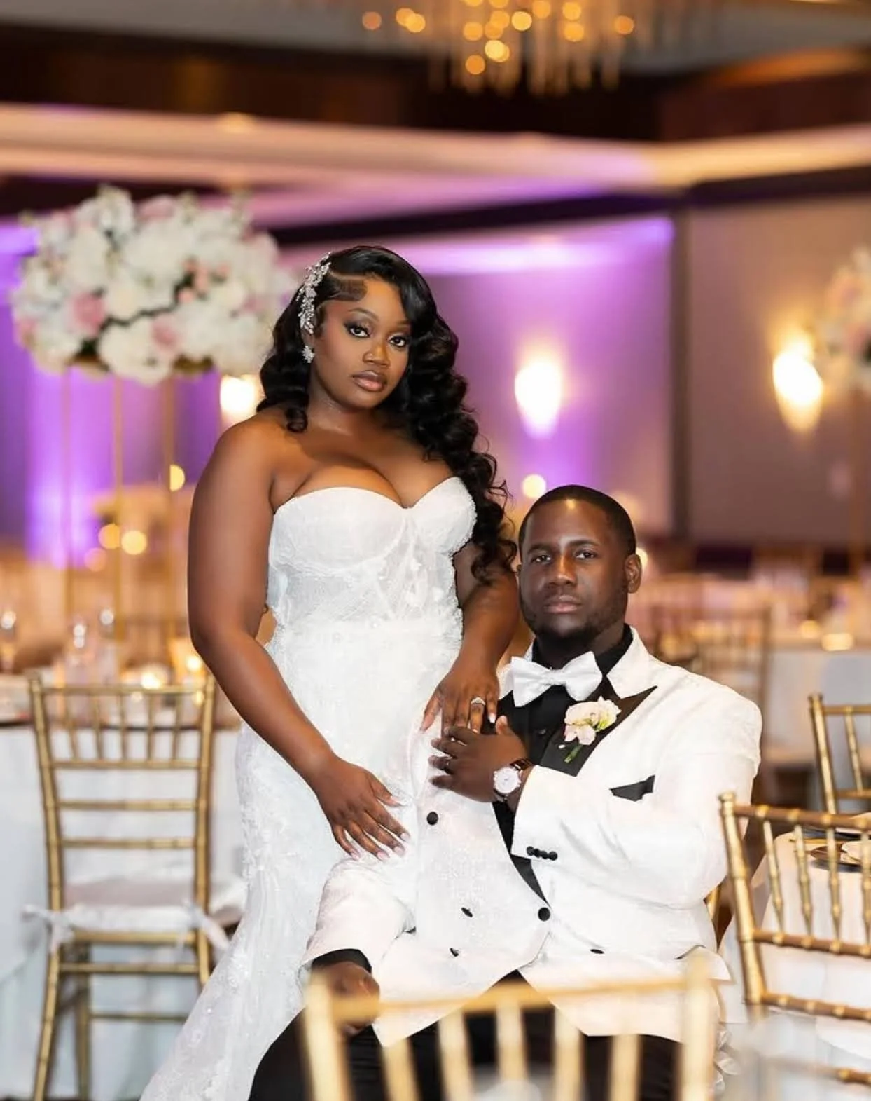 A couple on their wedding day inside their venue. The groom is sitting with the bride standing next to him.