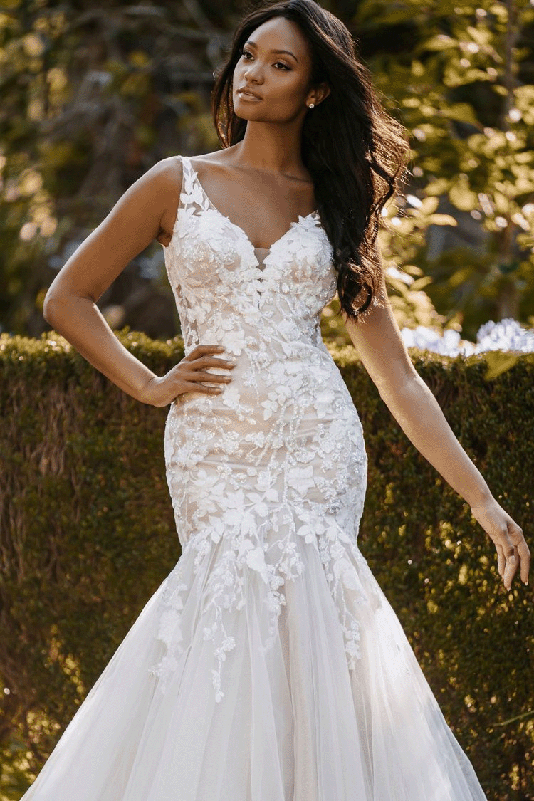 A woman in a white lace wedding dress standing outdoors with trees and bushes in the background.