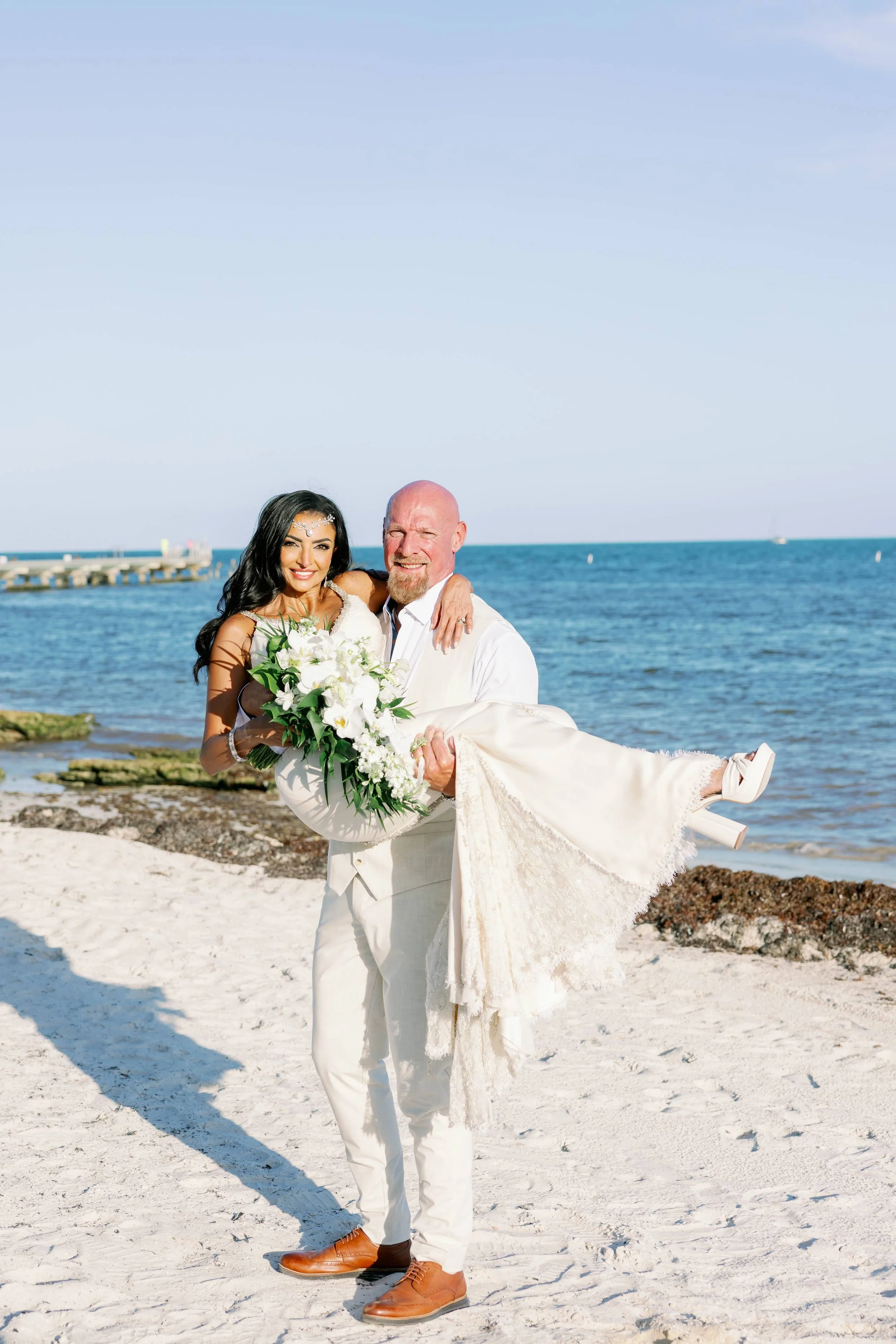 A couple on their wedding day on the beach. The groom is holding the bride while the bride holds a bouquet.