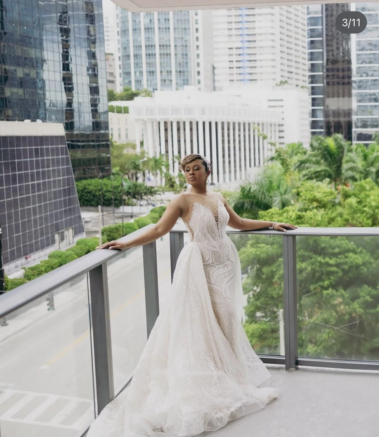 A woman in a white wedding dress standing on a balcony with city buildings and green trees in the background.