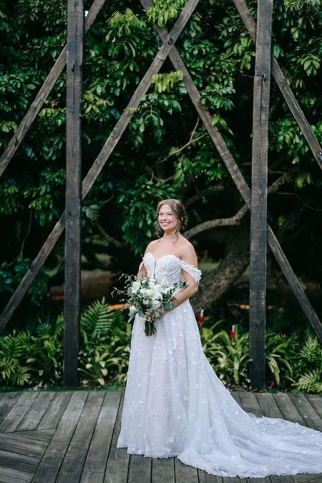 A bride smiling holding a bouquet outside in front of greenery.