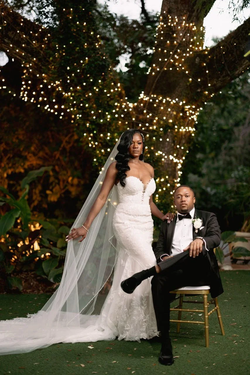 A couple on their wedding day in front of a tree with lights wrapped around the trunk. The groom is sitting on a chair while the bride stands next to him.