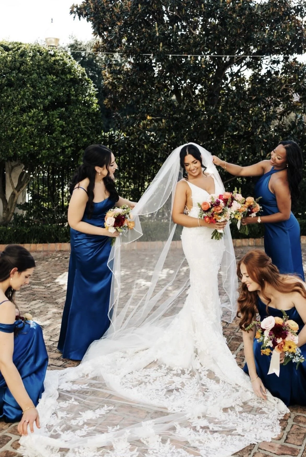 A bride smiling outside holding a color bouquet while her bridesmaids stand around her adjusting her veil and train.