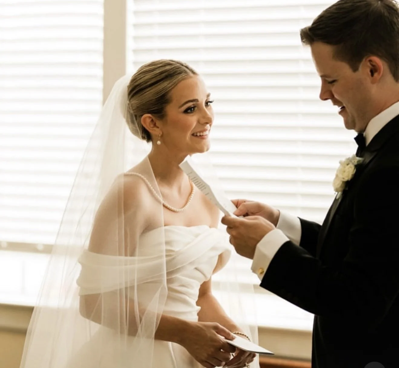 A bride and groom stand facing each other during their wedding ceremony, with the bride smiling and looking at the groom, who is reading vows from a piece of paper.