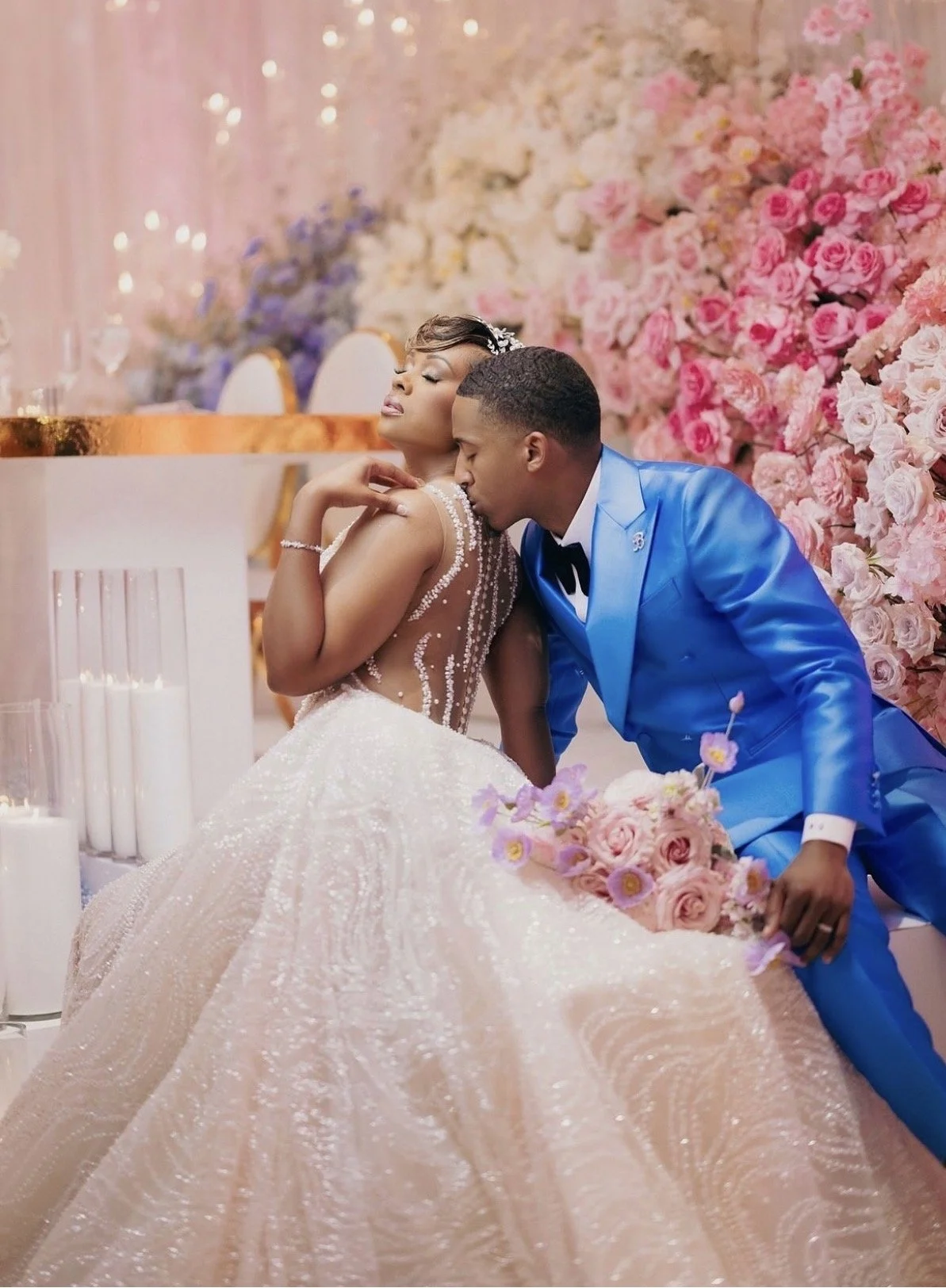 A couple on their wedding day in front of pink and purple flowers and the groom is kissing the bride's shoulder.