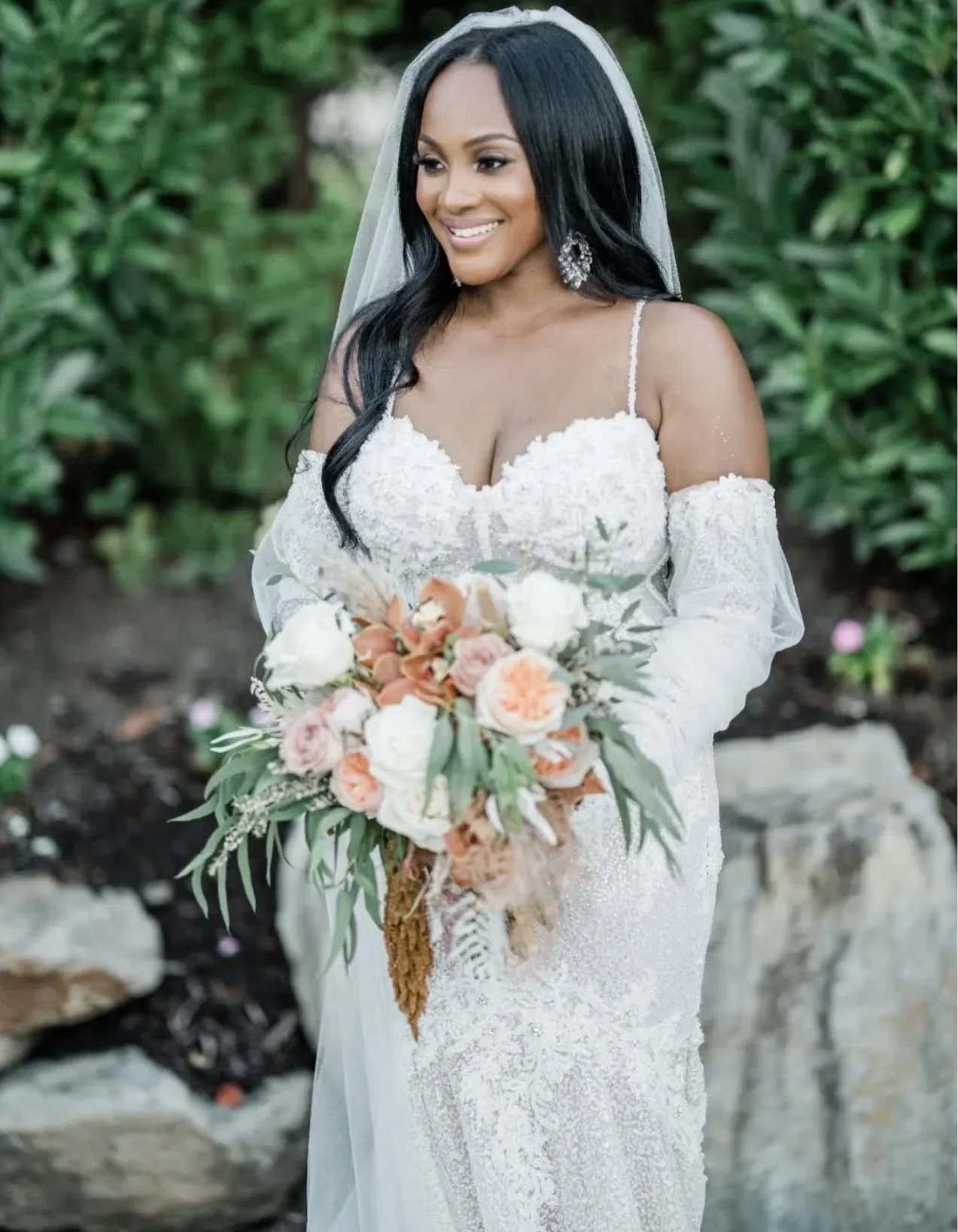 A bride with long black hair wearing a white lace wedding dress with off-shoulder sleeves, holding a bouquet of peach, white, and pink flowers, standing outdoors with greenery in the background.