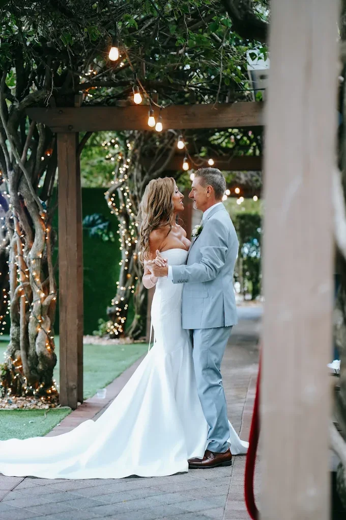 A bride and groom dance outdoors under string lights, surrounded by trees decorated with fairy lights, during their wedding celebration.