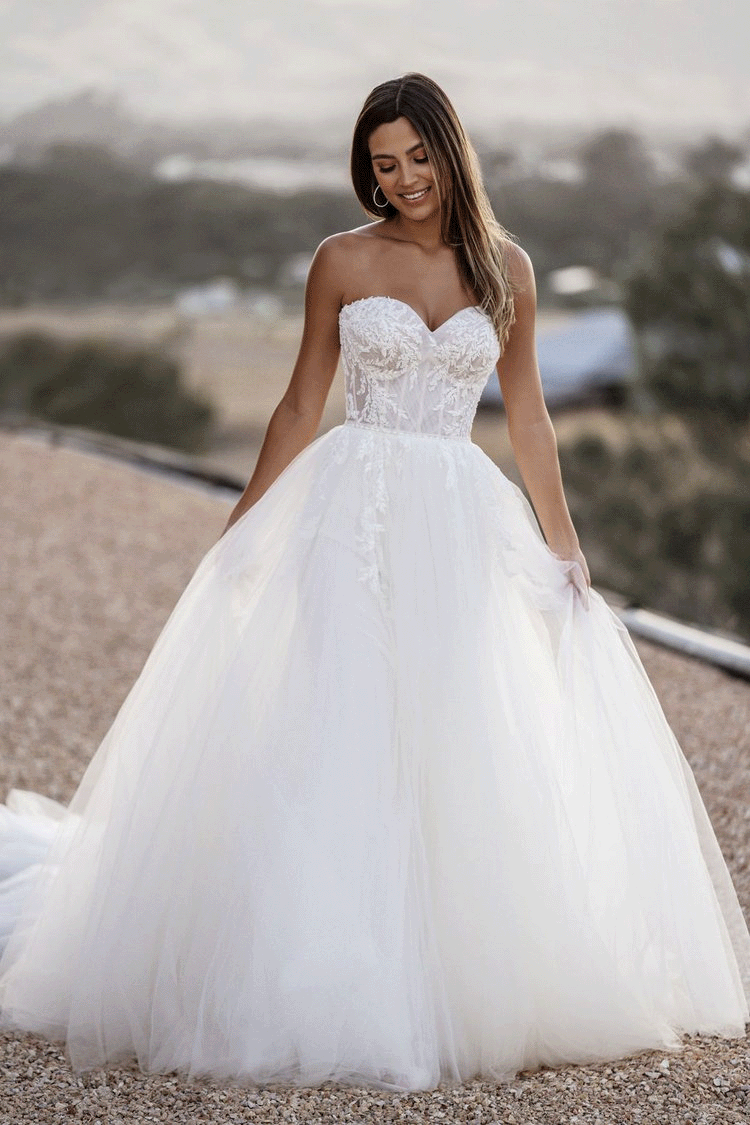 A woman in a strapless white wedding gown with a lace bodice and full tulle skirt, standing outdoors on a gravel path with a blurred natural landscape in the background, smiling and looking down.