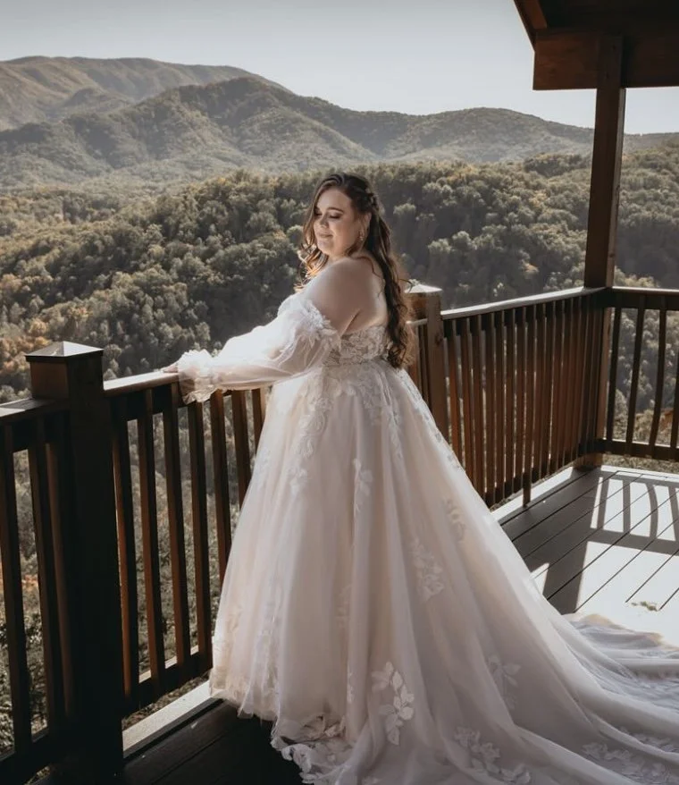 A woman in an elegant white wedding gown standing on a wooden balcony overlooking a mountainous landscape.