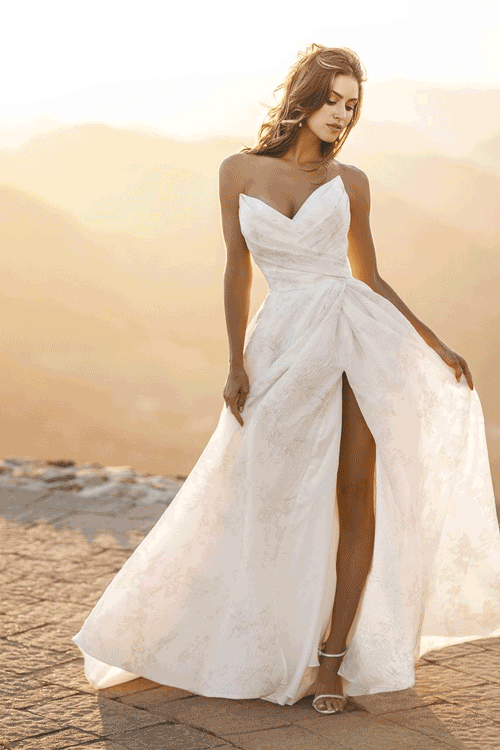 A woman in a strapless white wedding dress with a high slit, standing outdoors at sunset in a desert landscape.