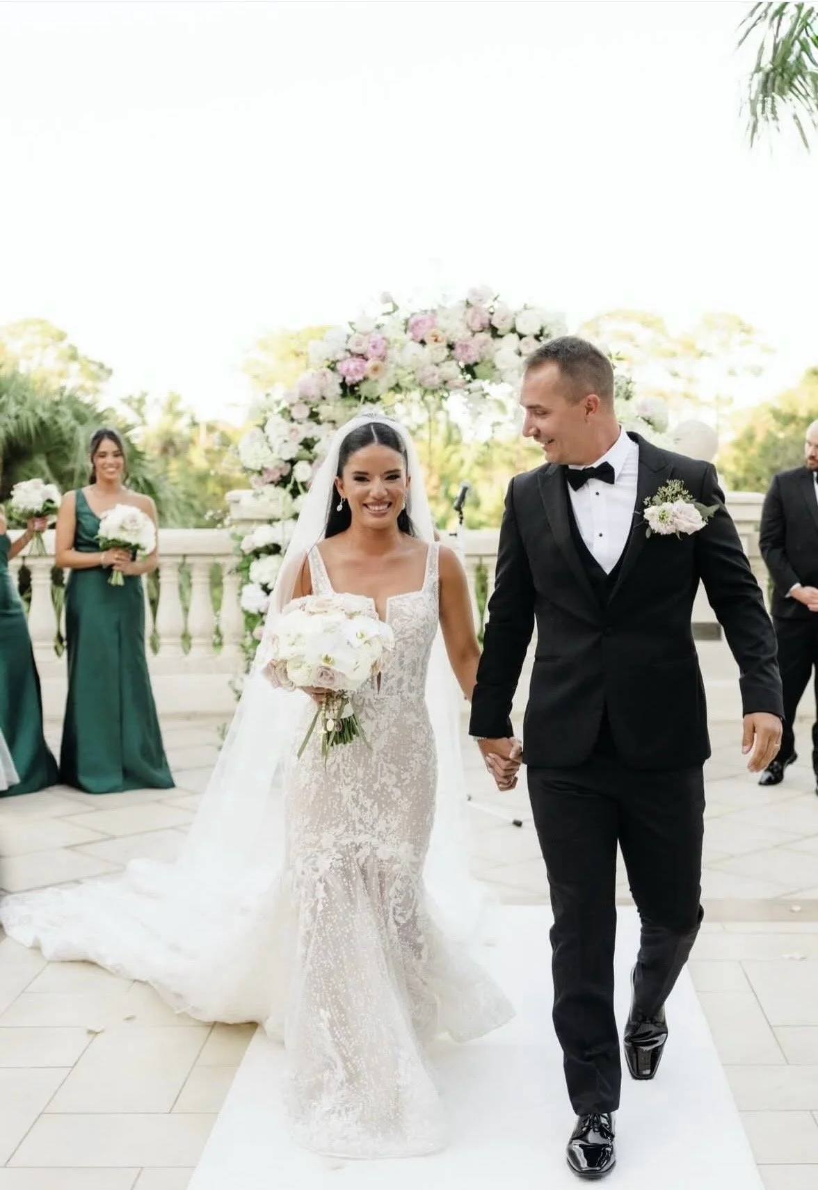 A bride and groom walking away from an arch of flowers while the bride is smiling and the groom is looking at her. 