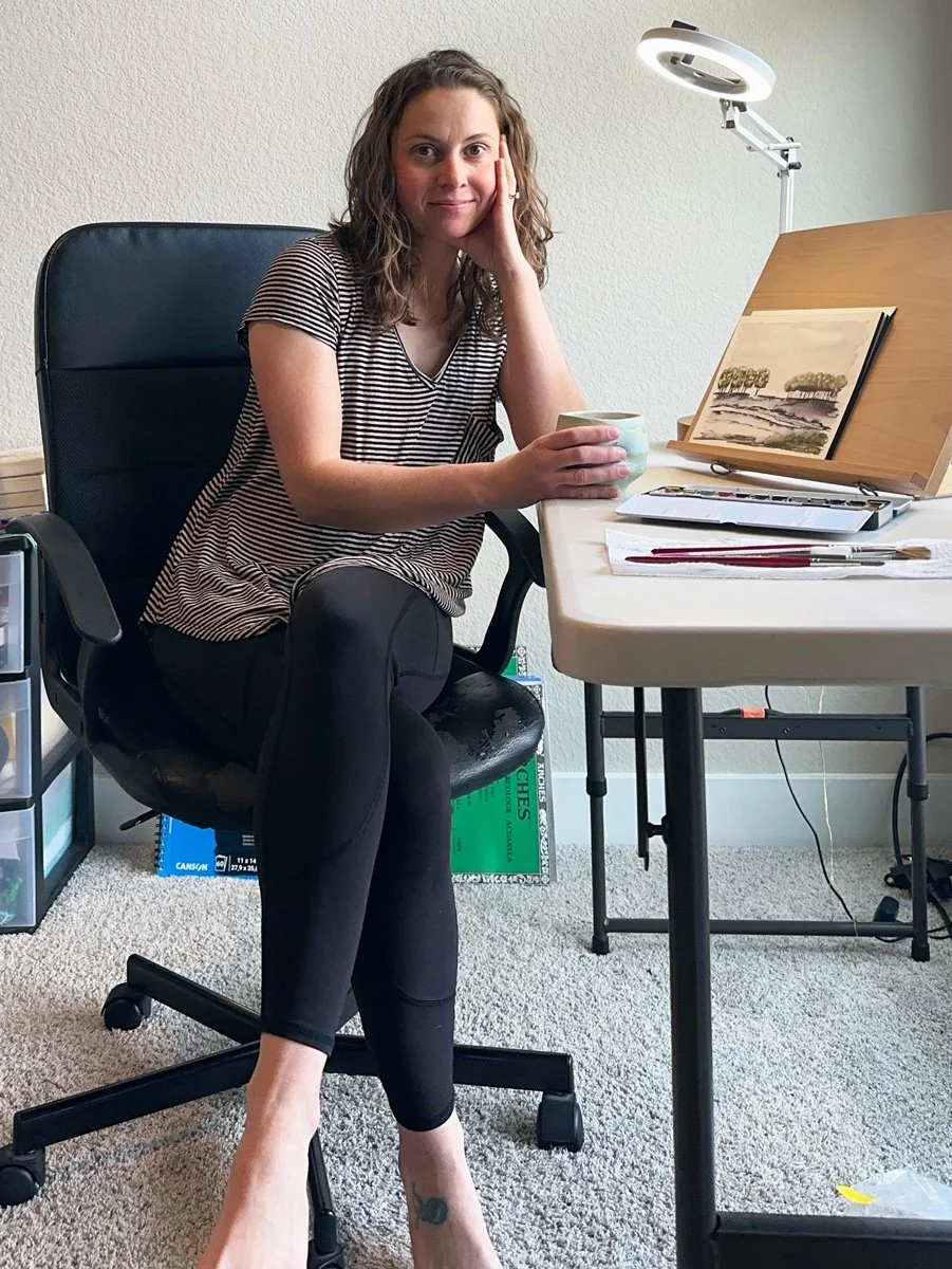 A woman with curly hair sitting on a black office chair at a desk, holding a mug, with her head resting on her hand, looking at the camera smiling. She is wearing a striped shirt and black leggings. The desk has a small landscape painting, brushes, and a laptop, with a lamp overhead. The room has a light-colored wall and carpeted floor.